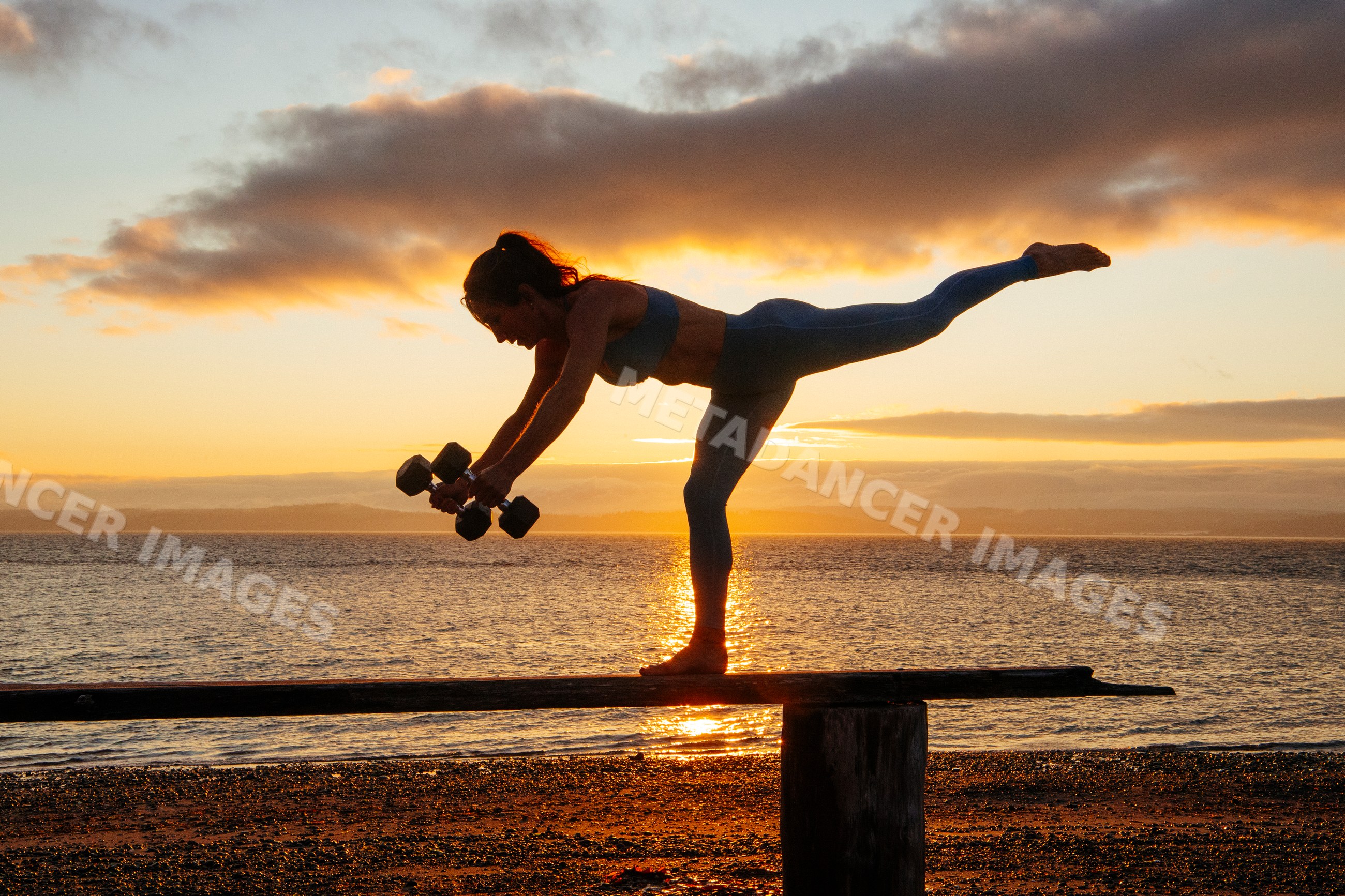 White woman balancing in yoga pose with dumbbells at sunset on beach