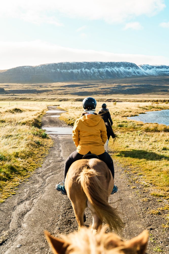 Two white people horseback riding on a trail beside a lake