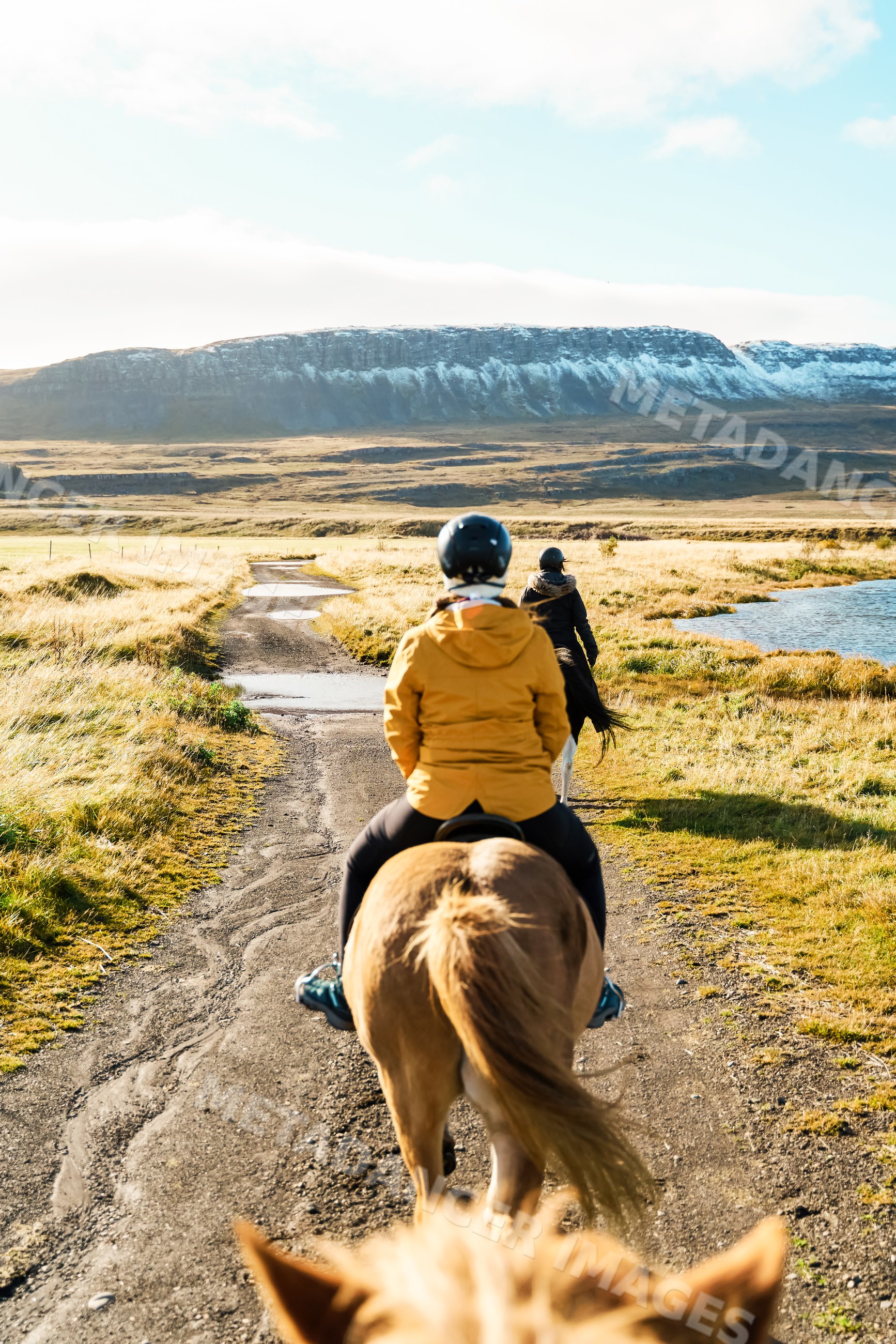 Two white people horseback riding on a trail beside a lake