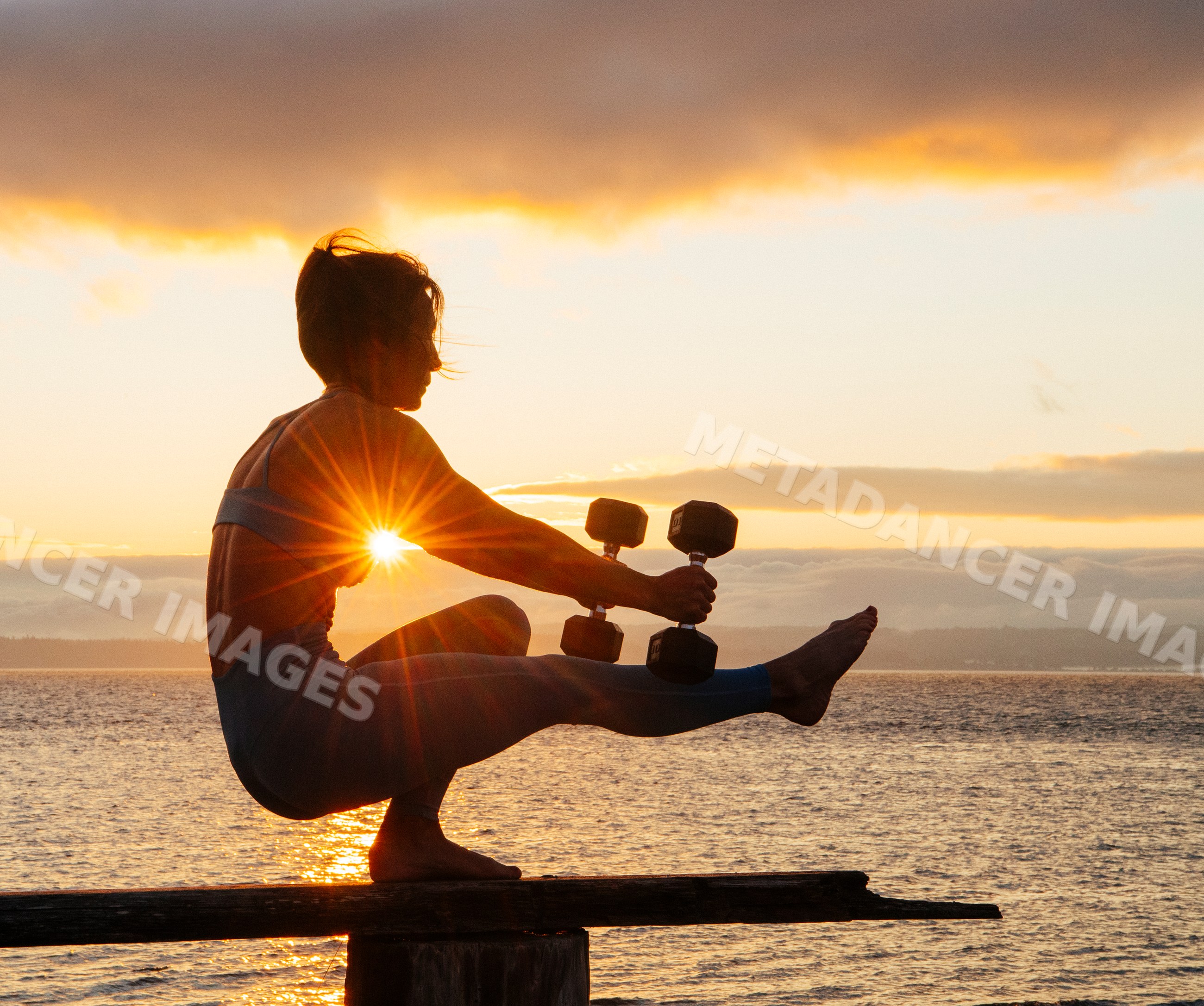 White woman balancing with dumbbells on wooden beam at sunset by water