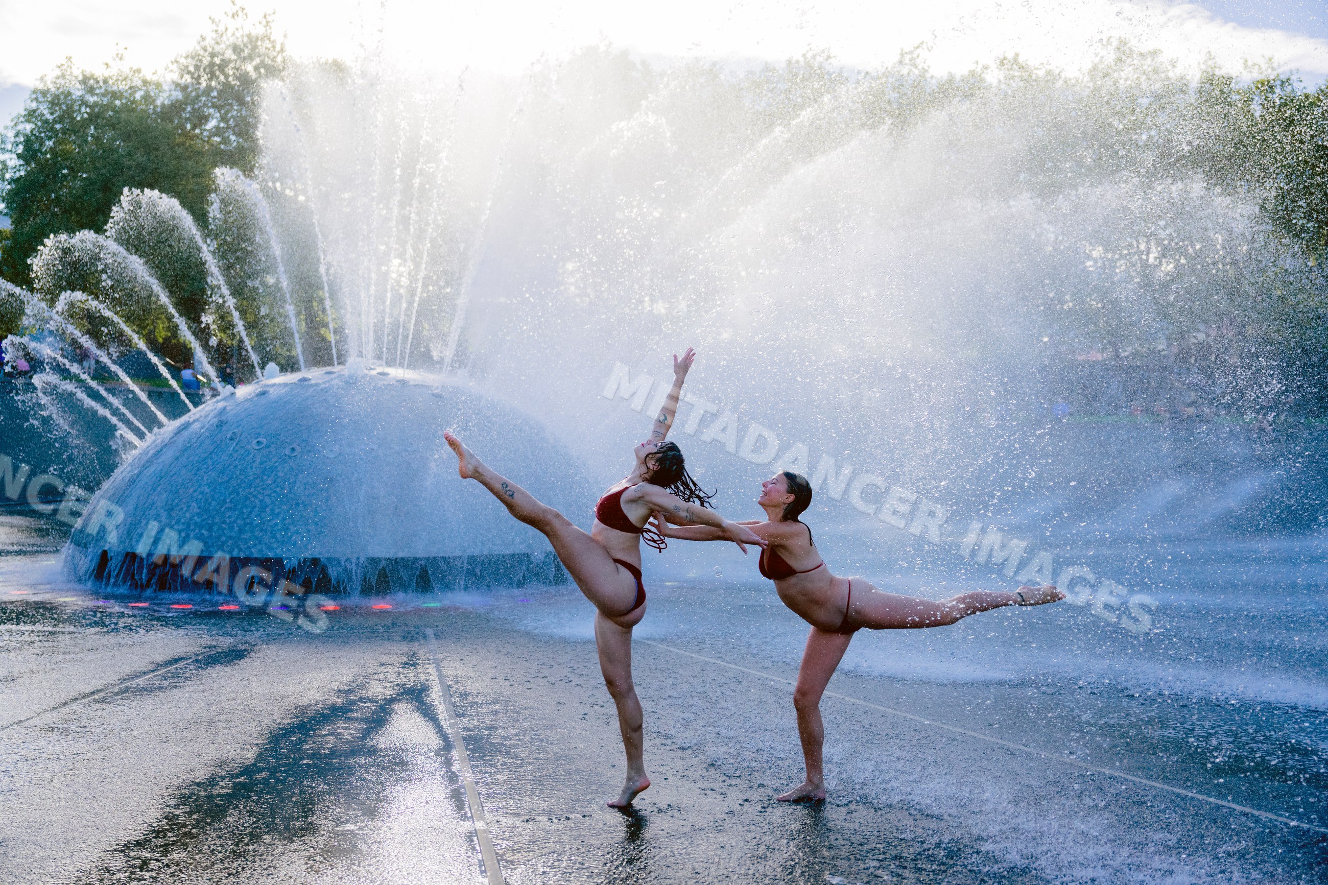 Two white women performing partner acrobatics yoga by water fountain