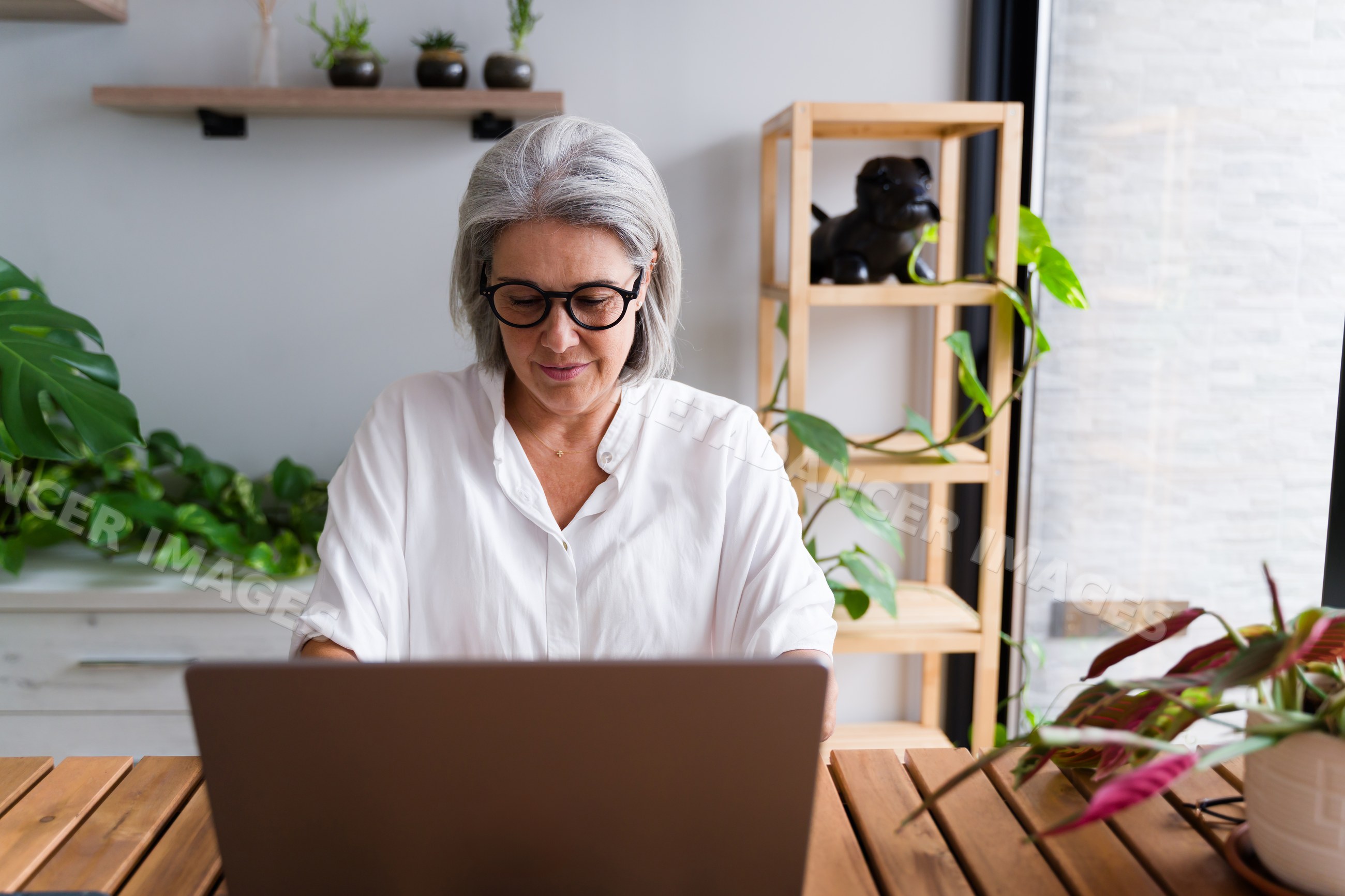 Senior white woman working on laptop at home office with plants