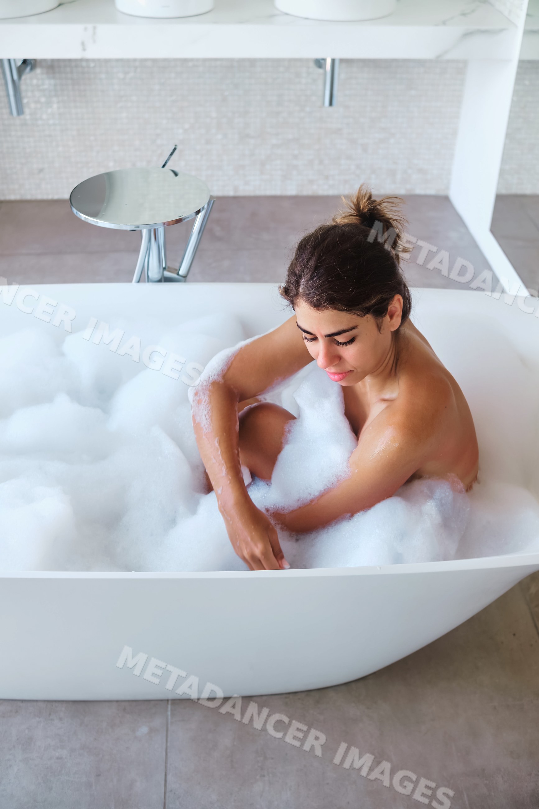 A white woman relaxes in a bubble bath in a modern bathroom