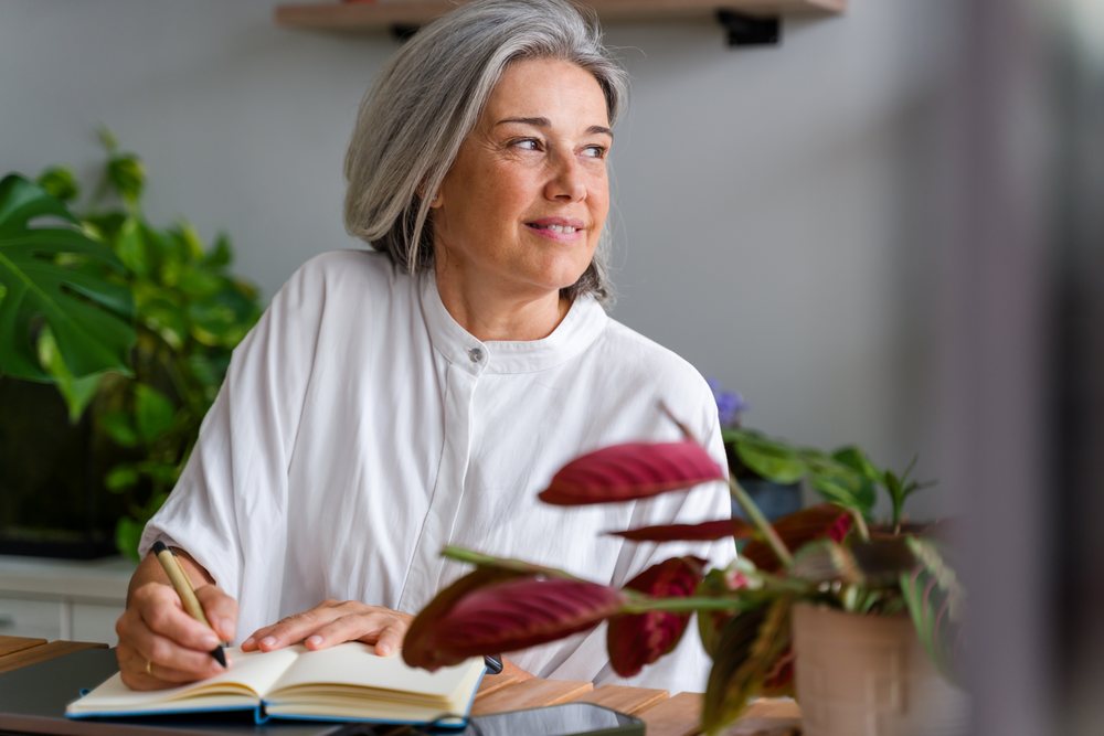 Mature white woman writing in notebook at home with indoor plants