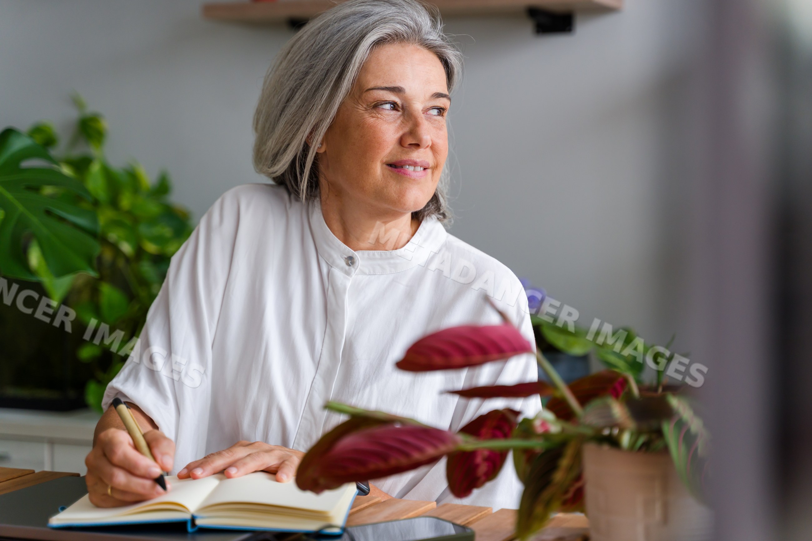 Mature white woman writing in notebook at home with indoor plants