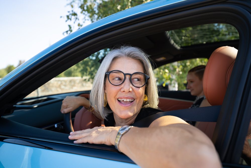 White woman smiling while driving a car with passenger in the backseat