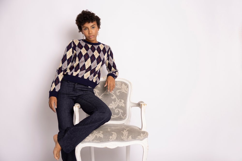 Hispanic woman in argyle sweater and blue jeans standing by armchair