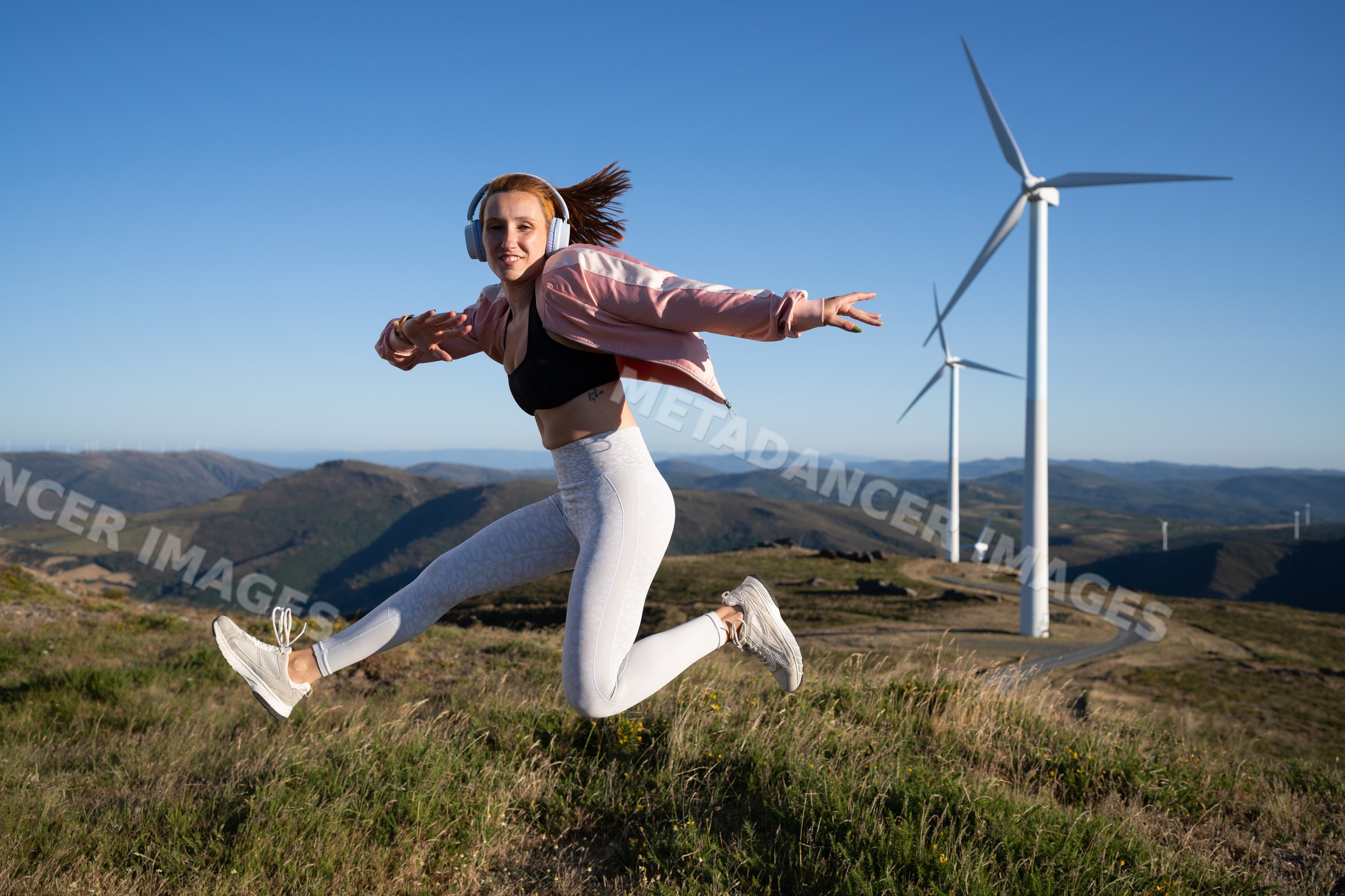 A young white woman jumps energetically outdoors near wind turbines