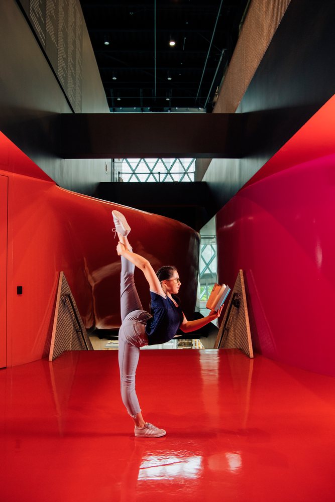A young woman reads a book while doing a yoga stretch in a library