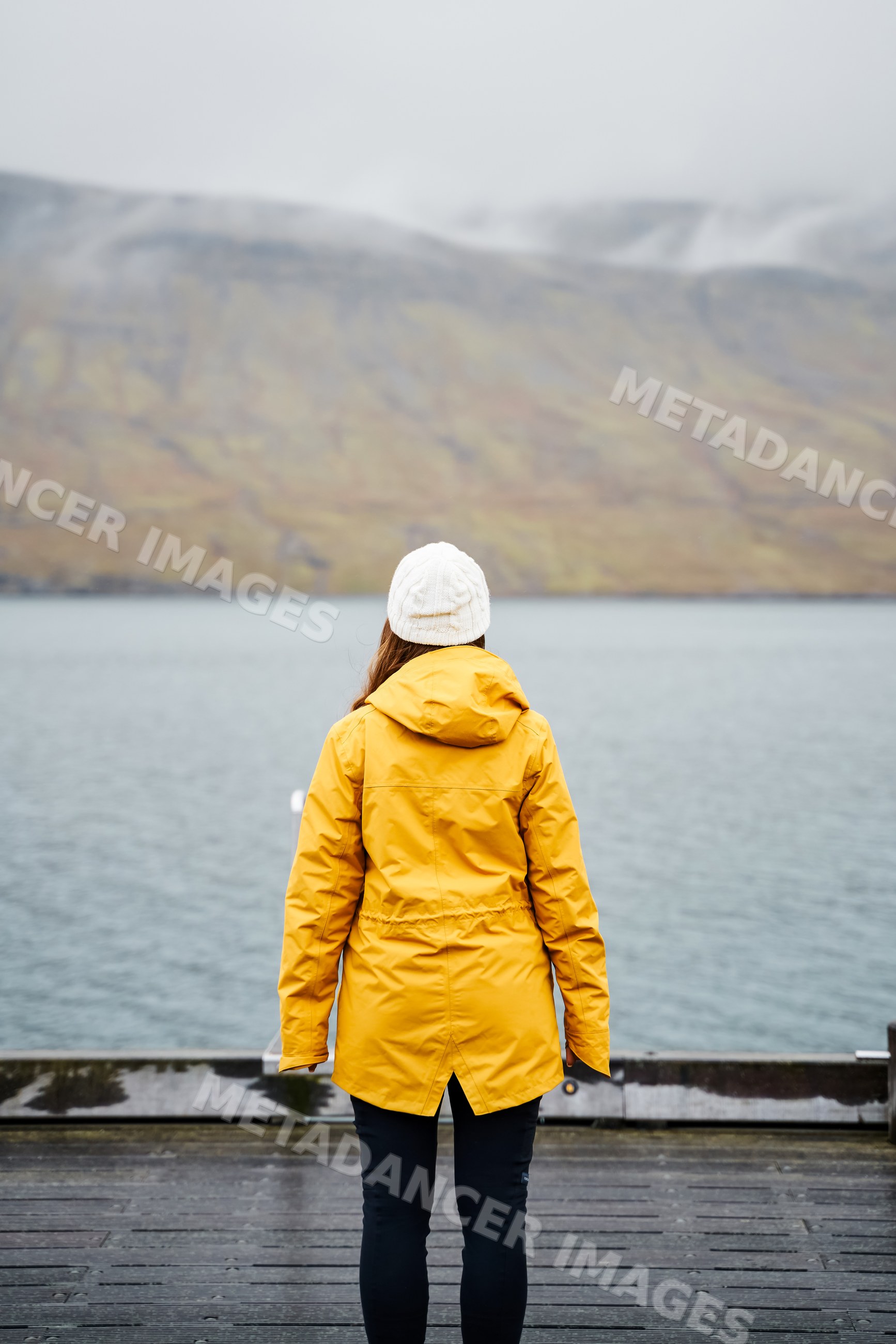 A white woman in a yellow jacket stands on a dock overlooking a misty