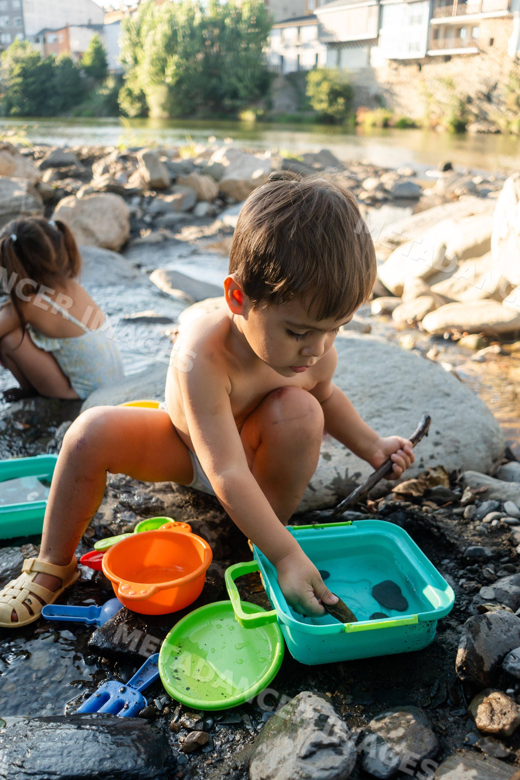 A young white boy playing with colorful toys by a rocky riverbank