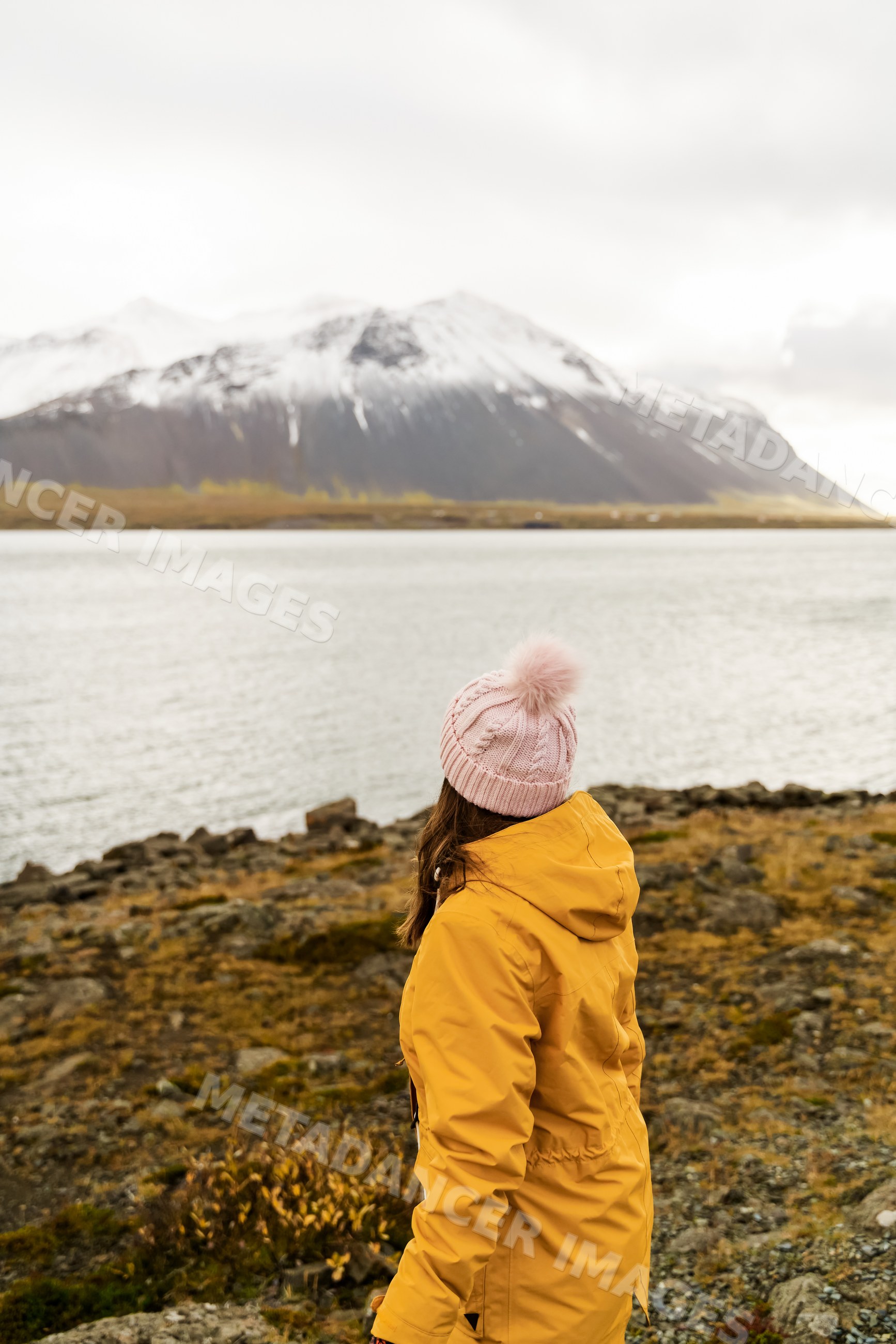 A white woman in a yellow jacket looks at a snowy mountain over a lake