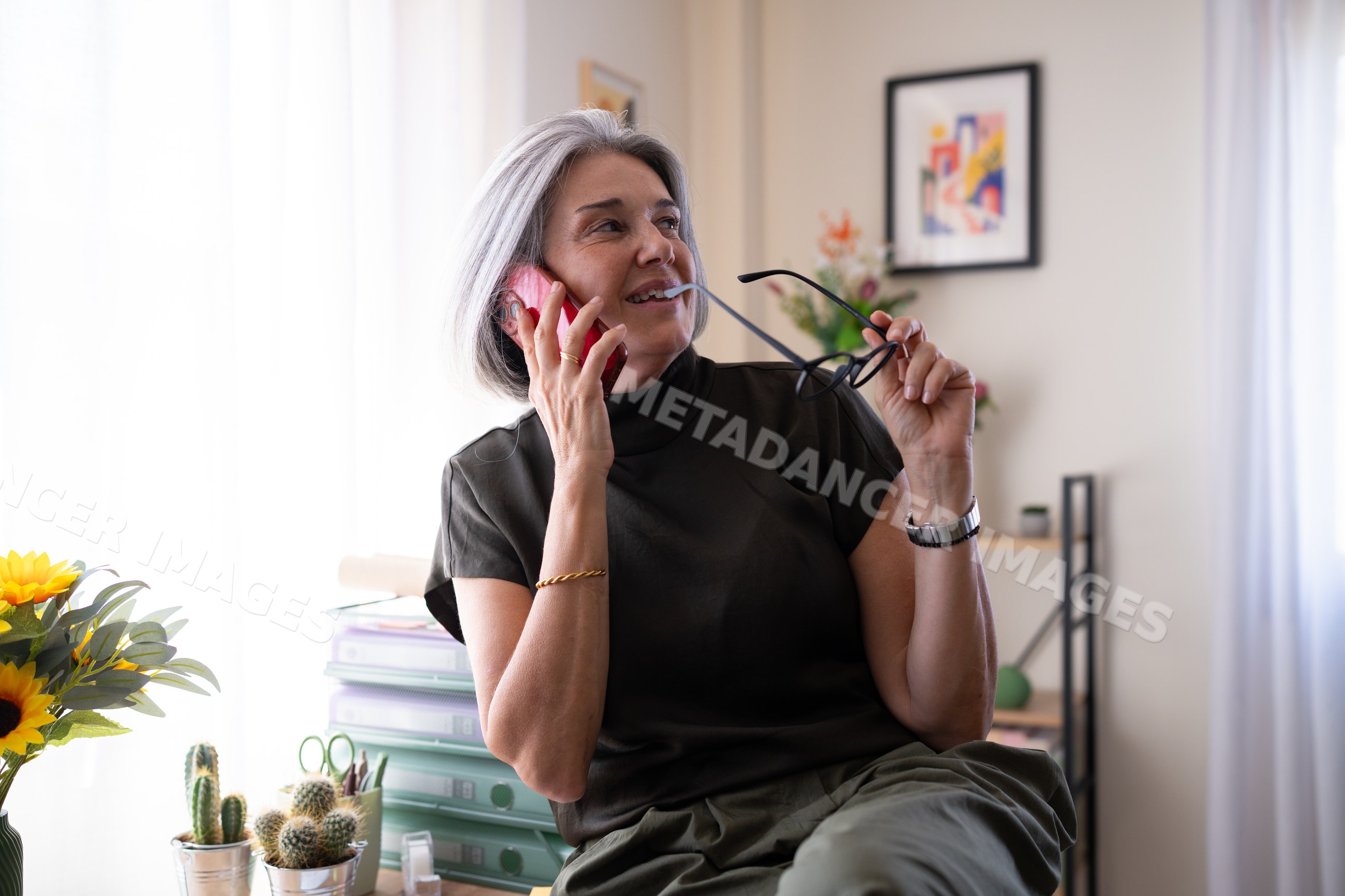 White woman talking on phone holding glasses in home office