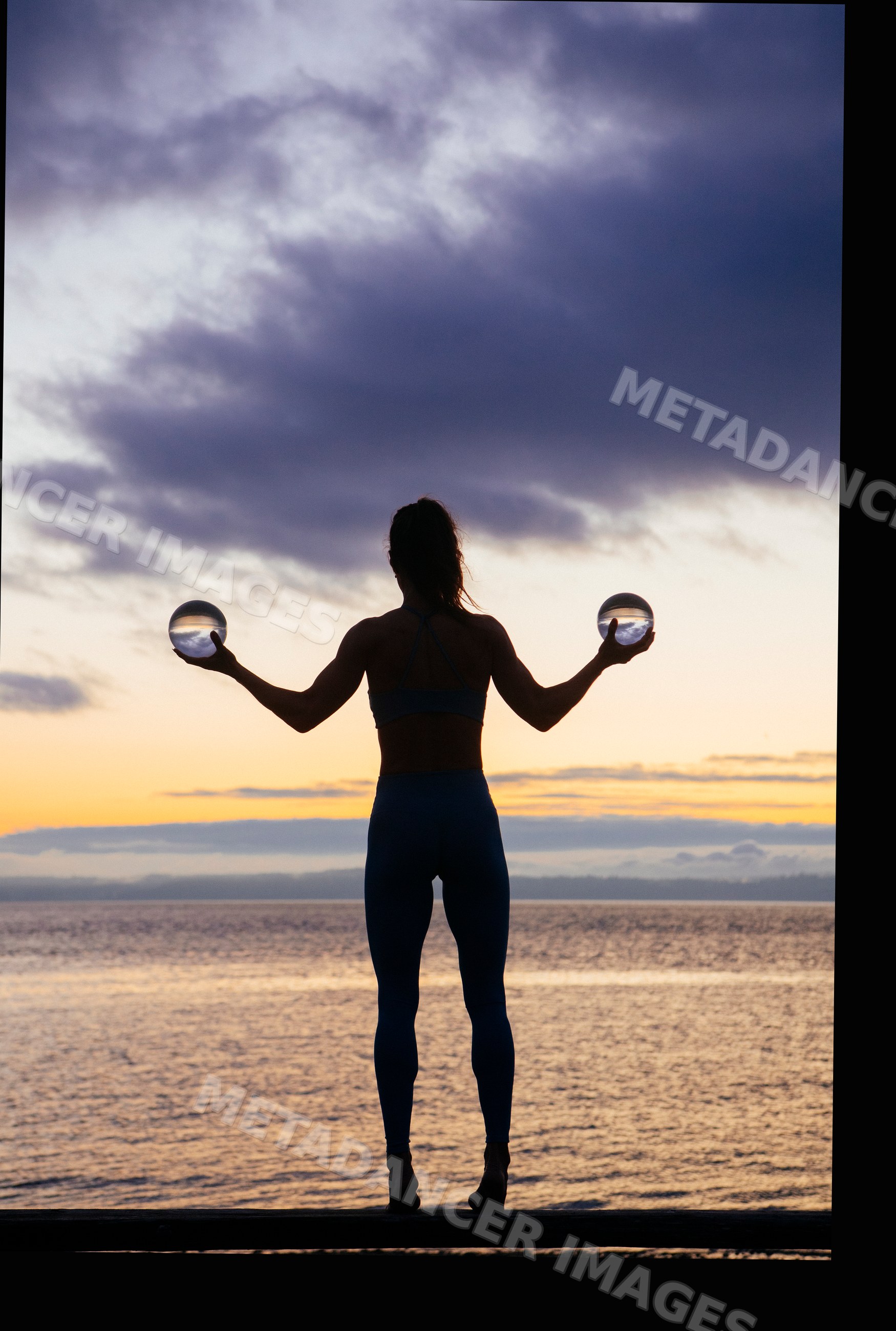 White woman silhouette holding crystal balls by waterfront at sunset