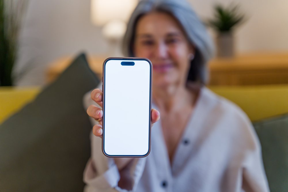 Senior white woman showing smartphone with blank screen indoors