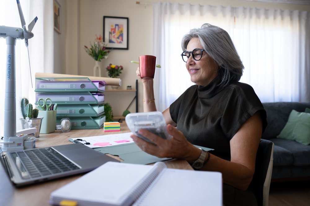 Senior white woman holding cup working at home office desk with laptop