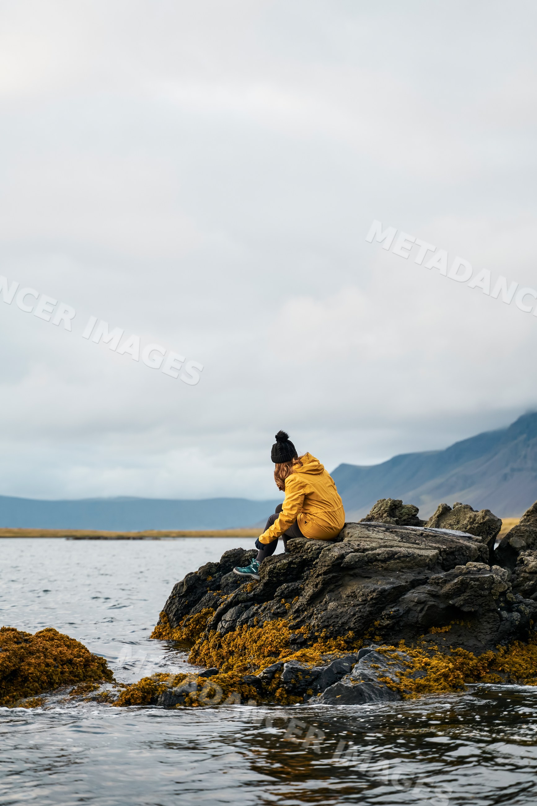 A white person sitting on seaweed-covered rocks by the sea
