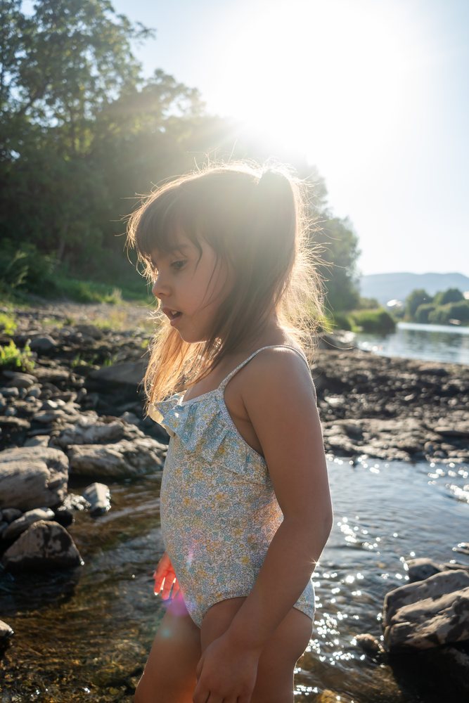 A white girl in a swimsuit explores rocky riverbank water on a sunny