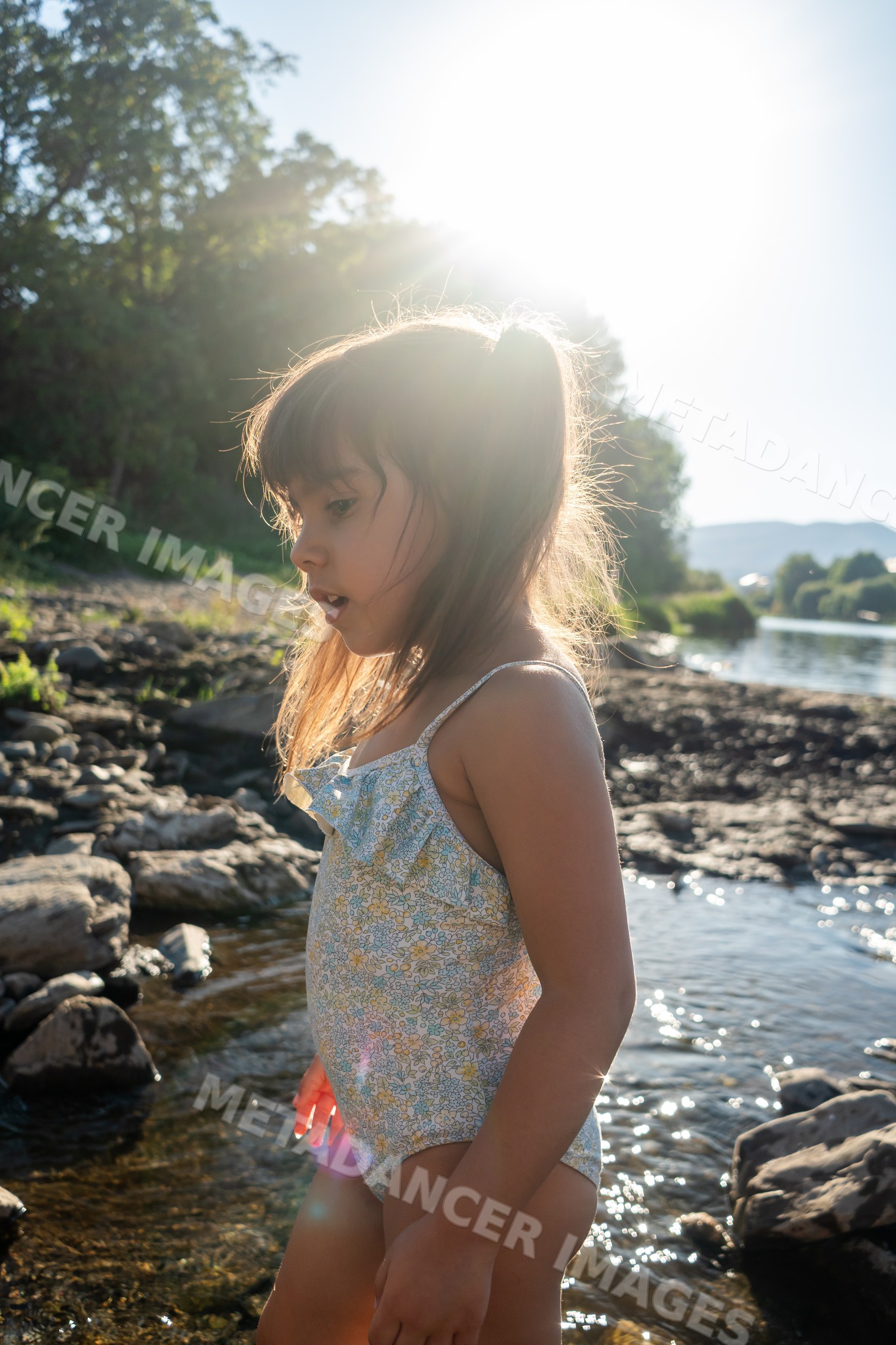 A white girl in a swimsuit explores rocky riverbank water on a sunny