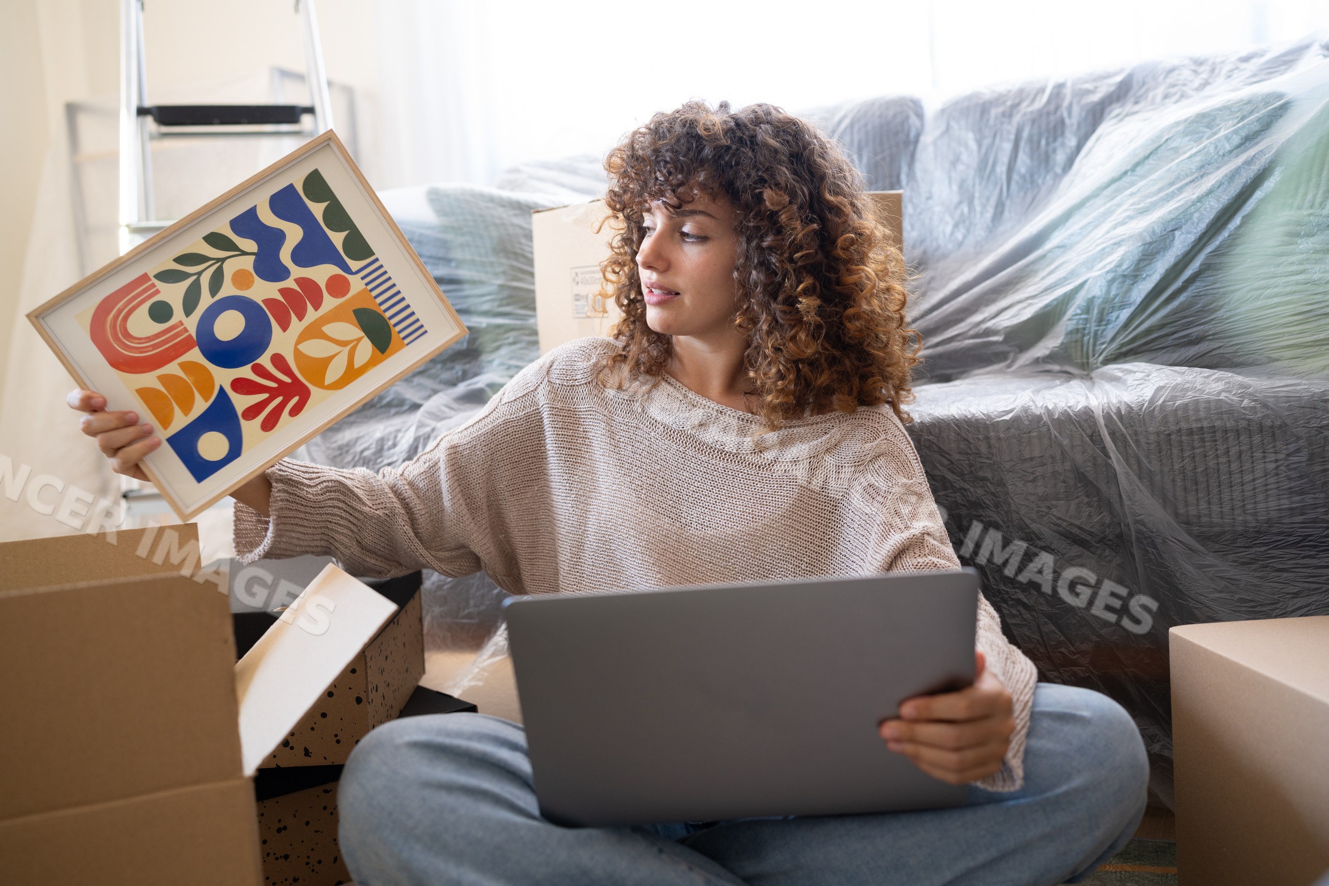 White woman holding artwork and laptop surrounded by moving boxes