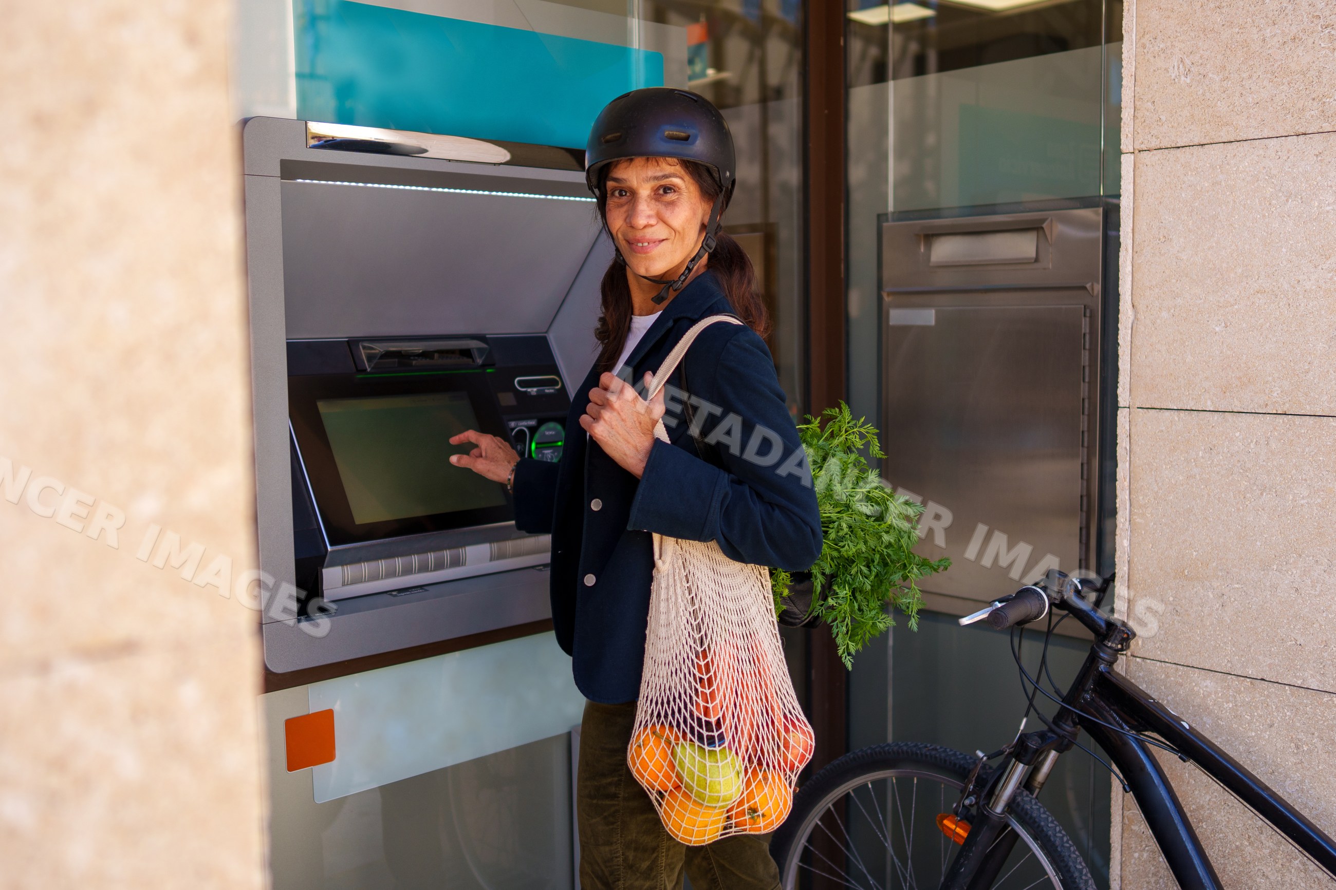 Hispanic woman using atm with reusable bag of fresh produce by bicycle