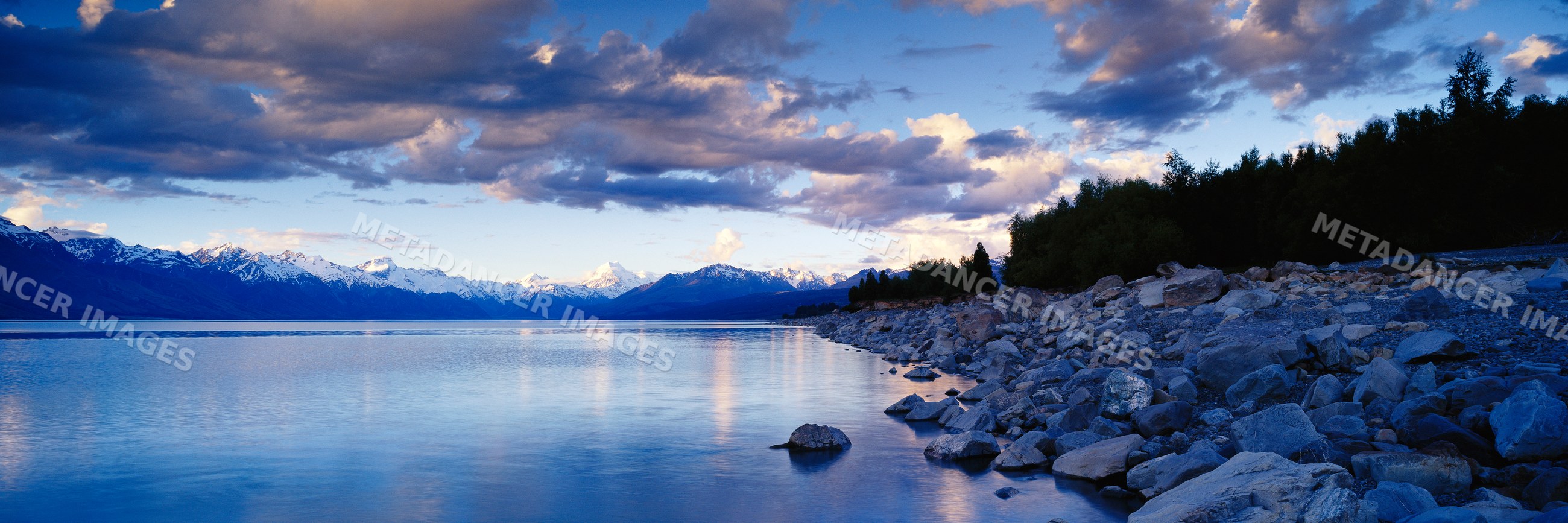 Lake with rocky shoreline and snow-capped mountains under twilight