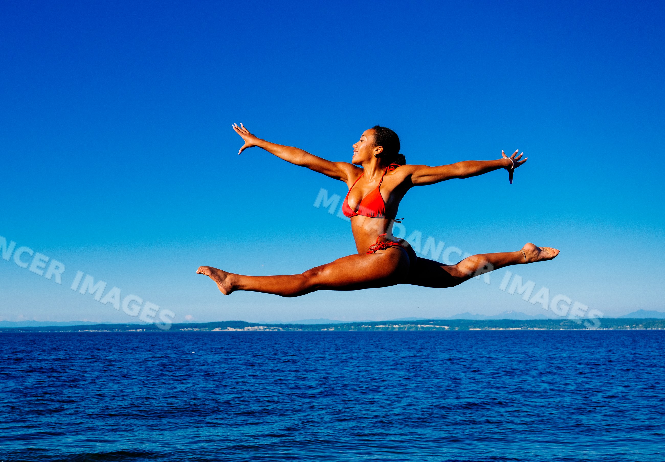 A woman in a red bikini jumps in a split over the ocean in WA, USA