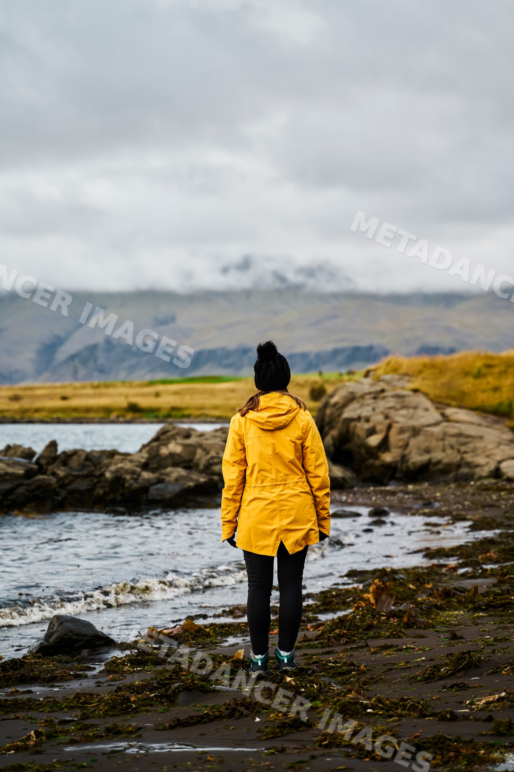 A white woman stands on a rocky shore with seaweed