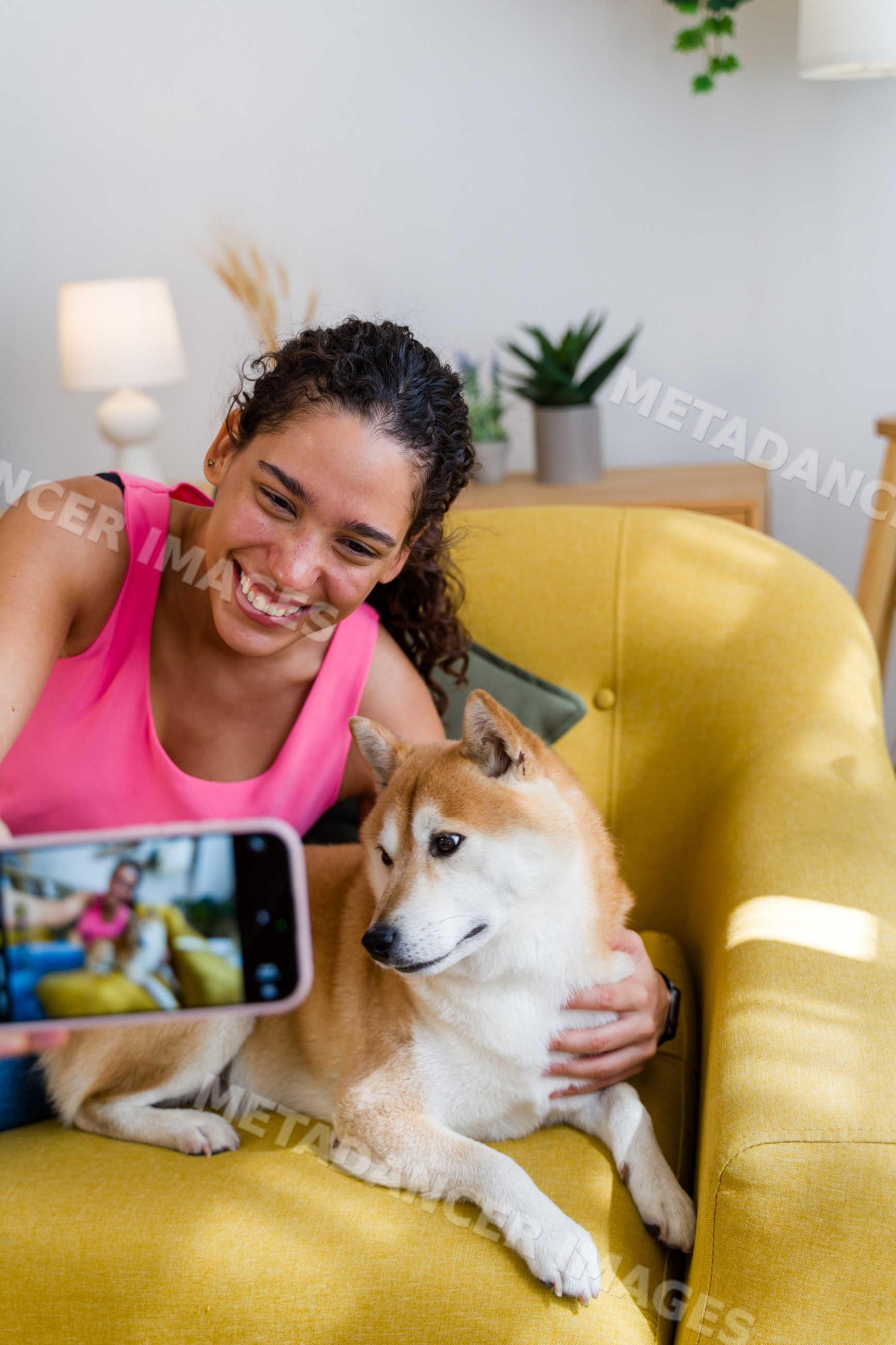 A Hispanic woman takes a selfie with her dog on a yellow armchair