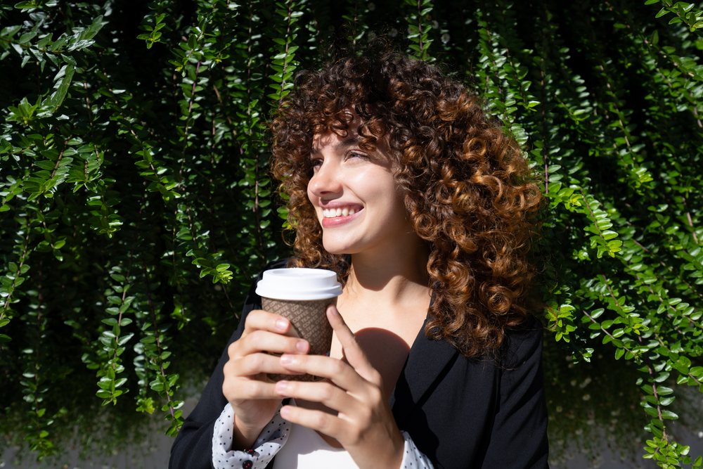 White woman enjoying coffee outdoors with curly hair and bright smile