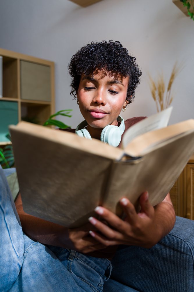 African American woman reading book indoors with headphones on sofa