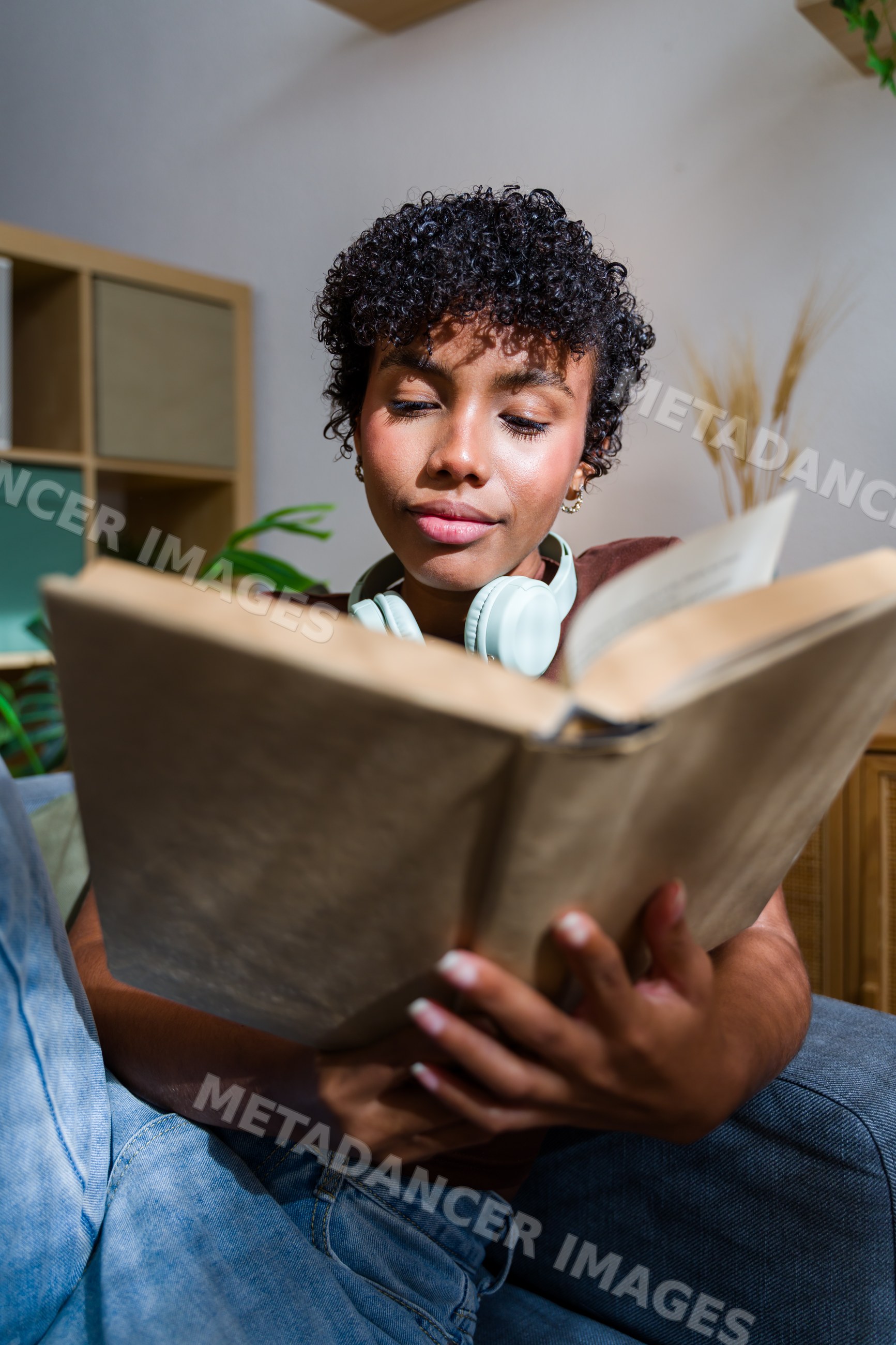 African American woman reading book indoors with headphones on sofa