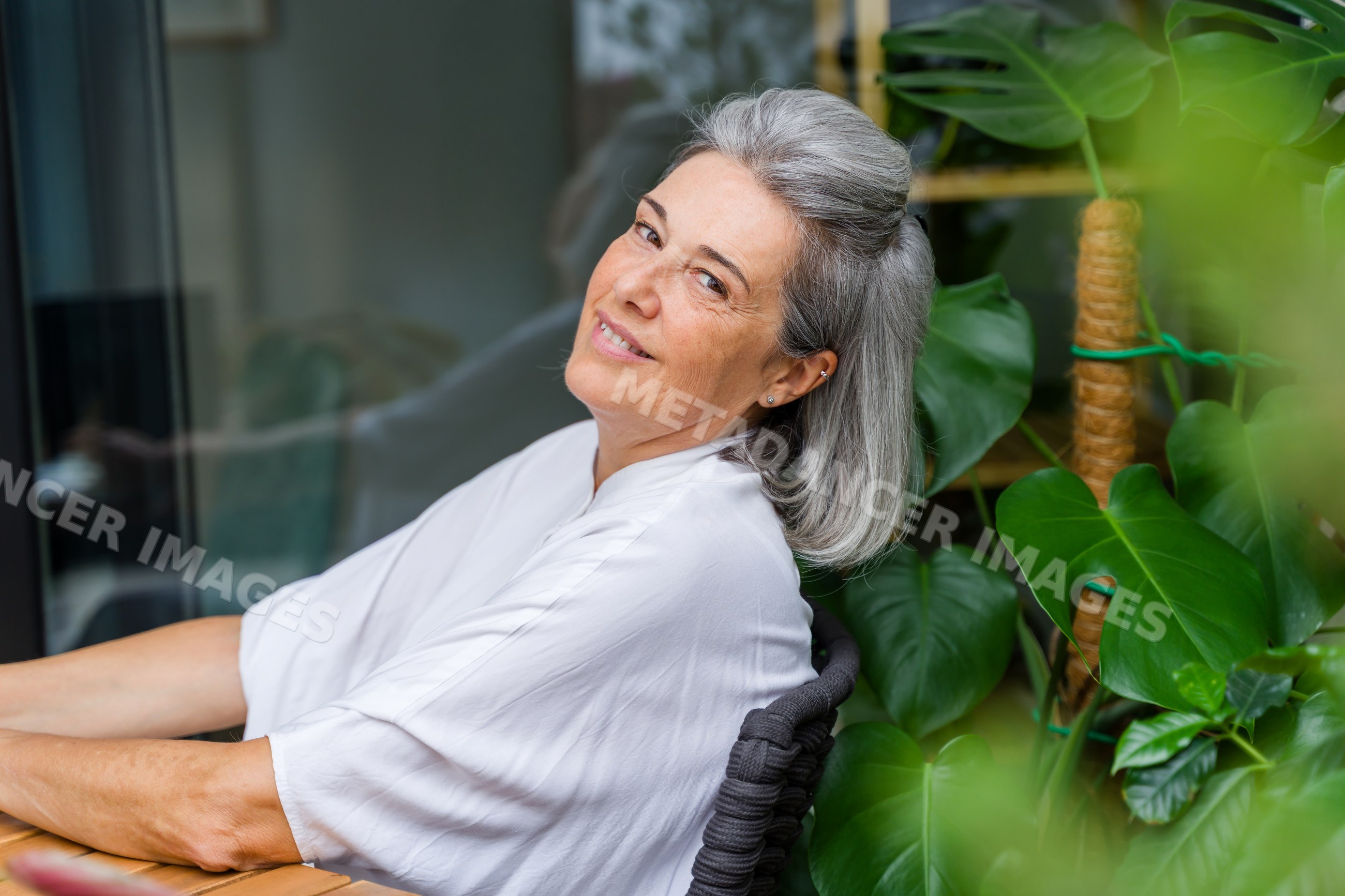 Smiling mature white woman sitting indoors with large leafy plant