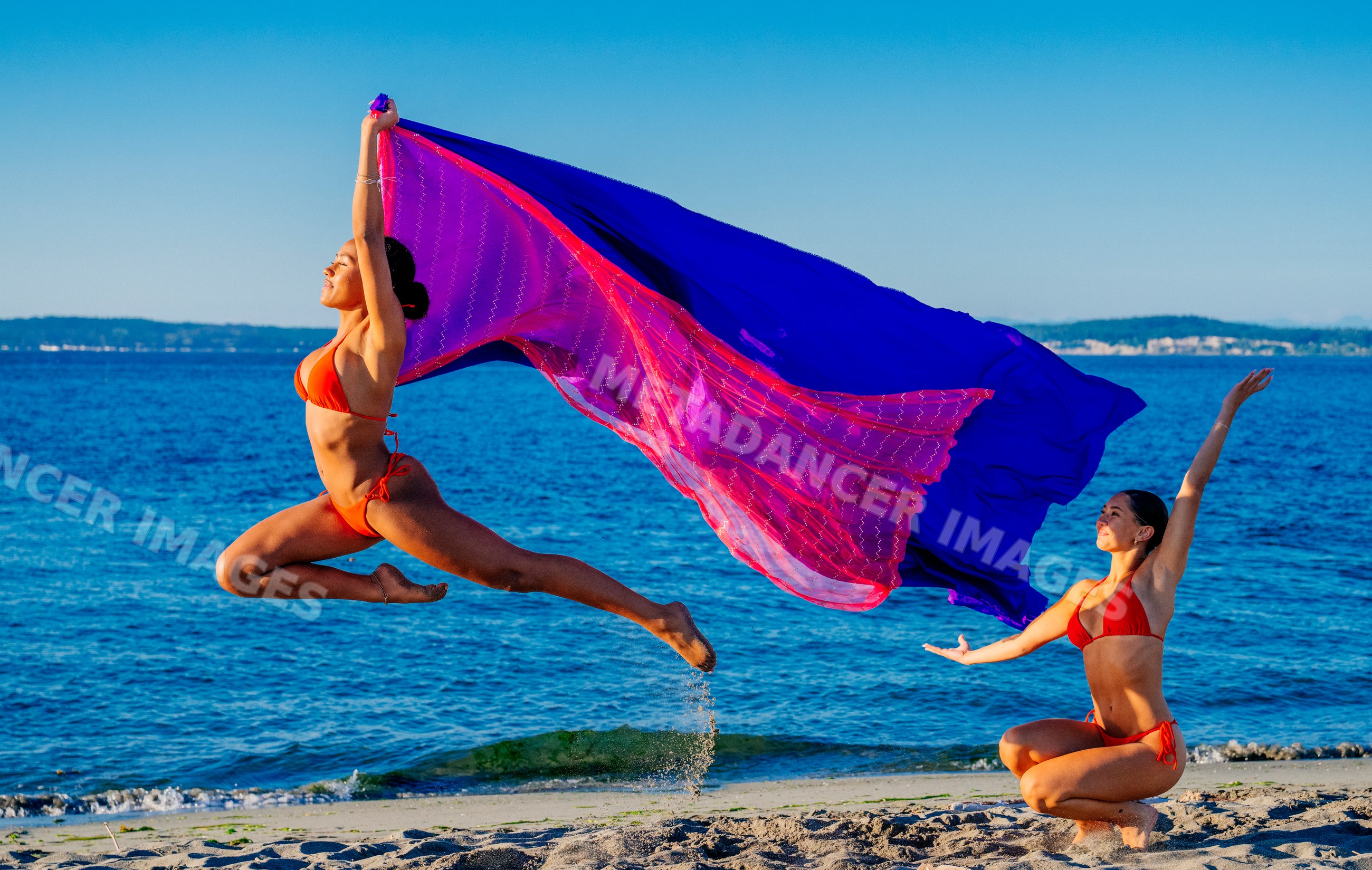 Two women perform partner yoga with flowing fabric on a beach