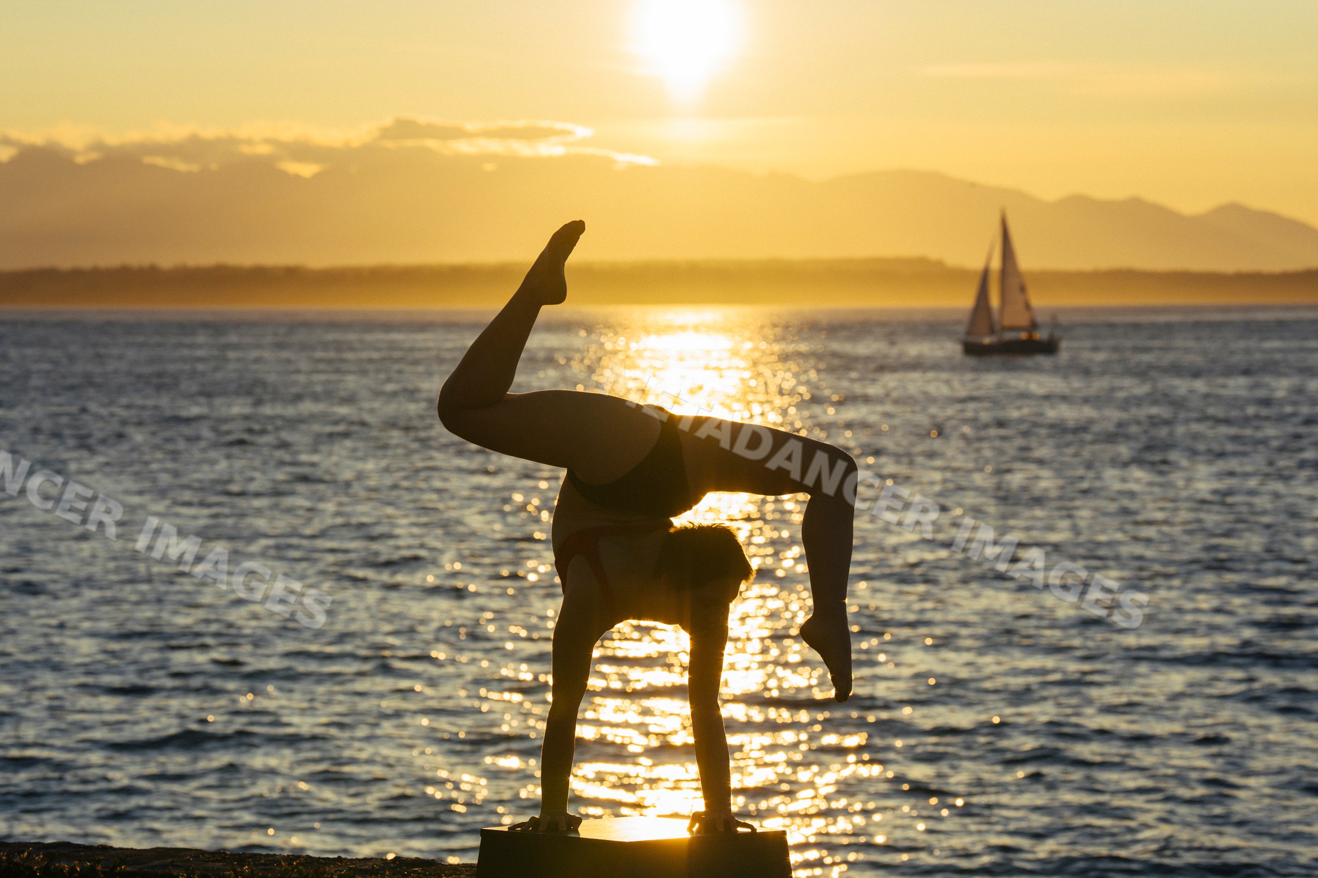 White person doing handstand by waterfront at sunset with sailboat