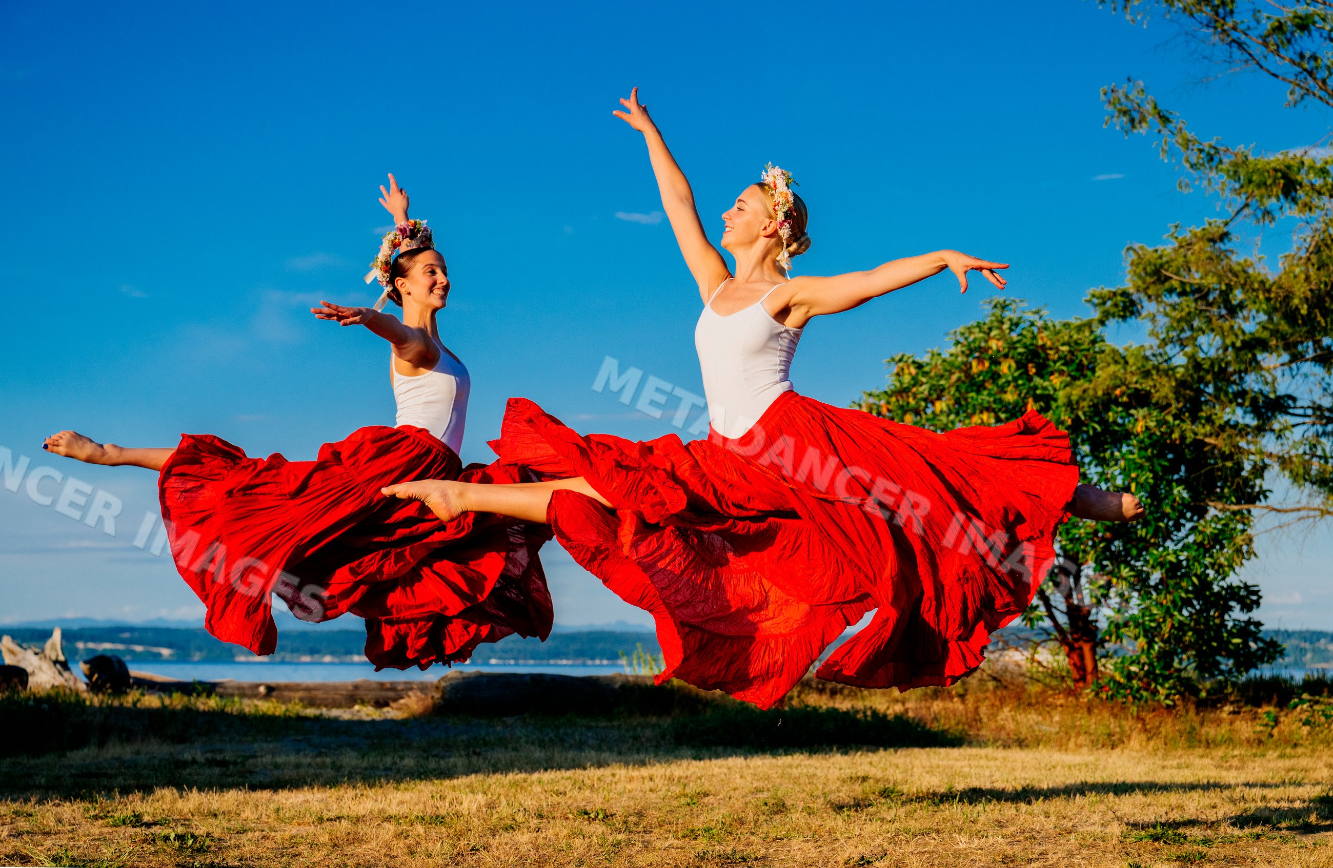 Two white women jump gracefully outdoors wearing red skirts and flower