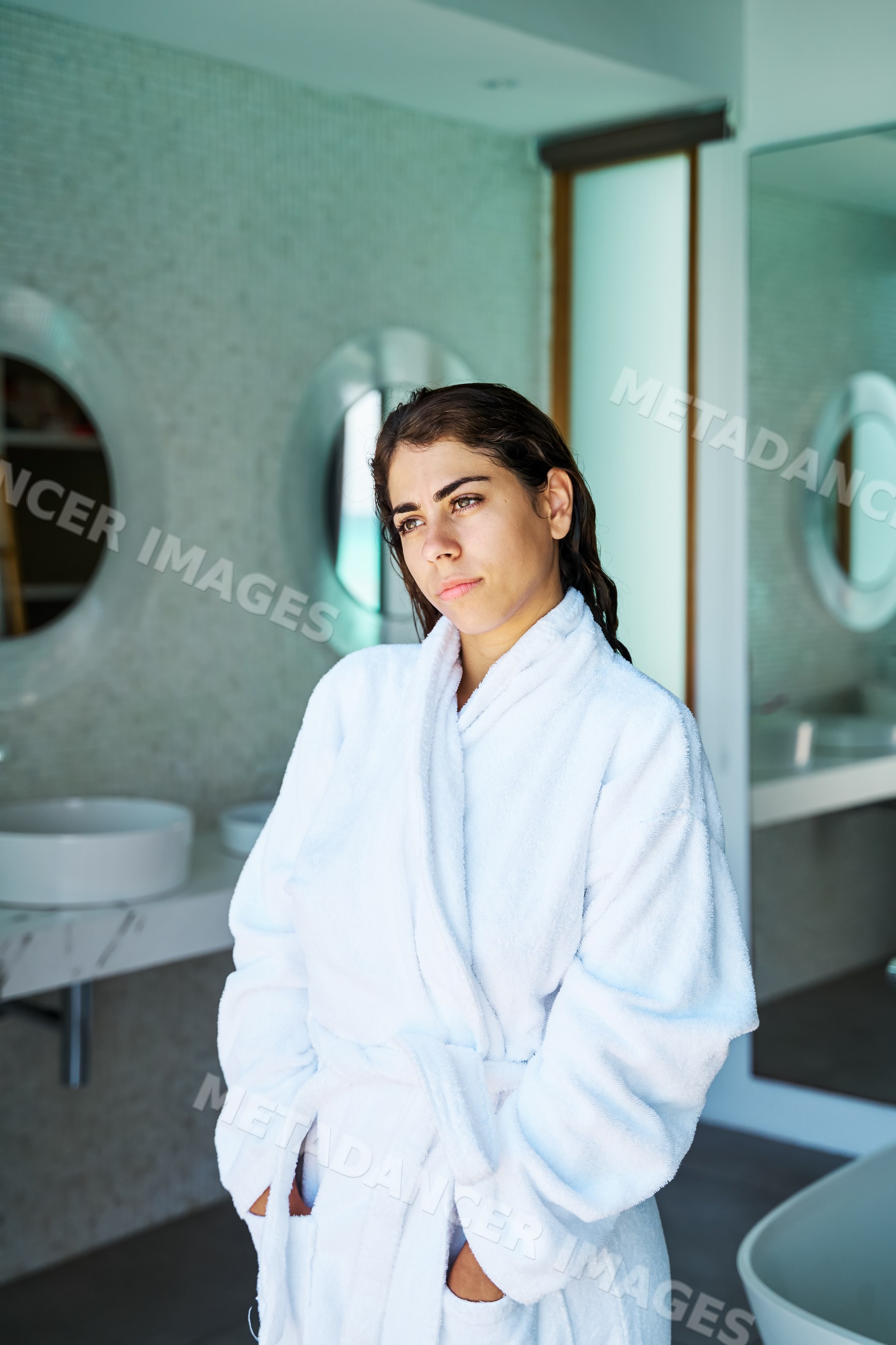A woman wearing a white bathrobe stands contemplatively in a bathroom