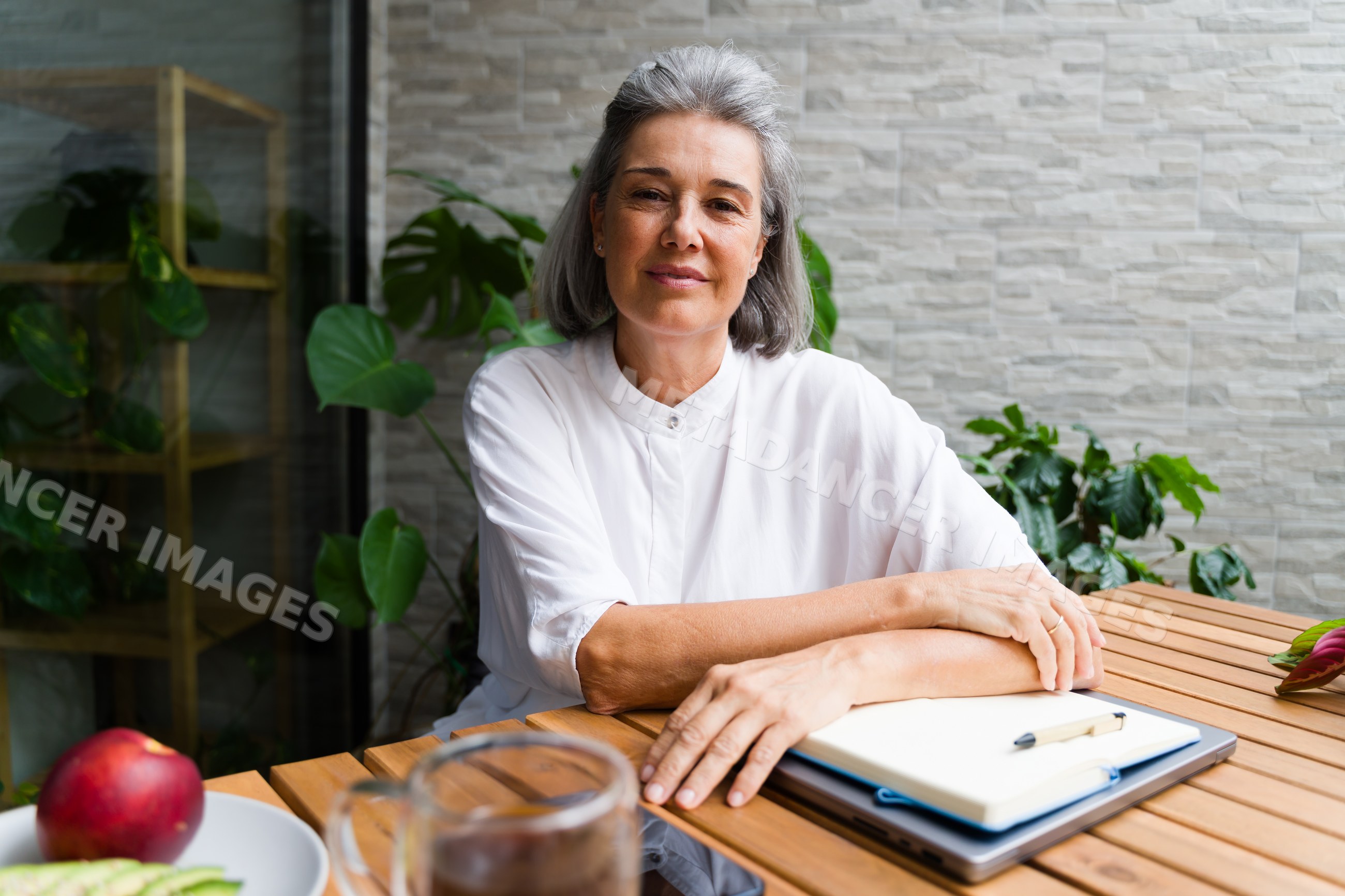 Mature white woman sitting at wooden table with notebook and plants