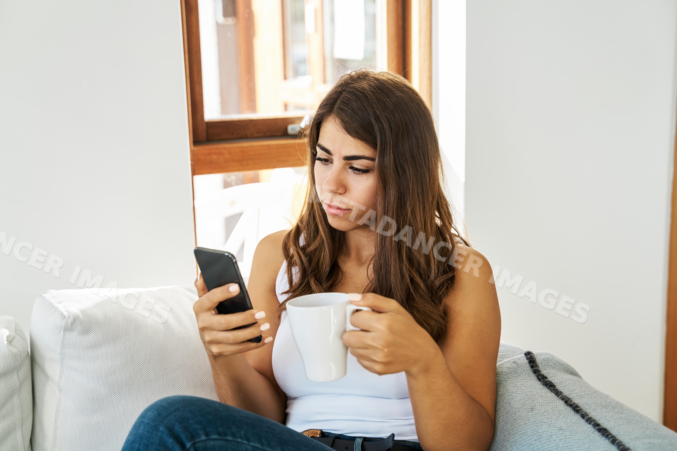 A white woman using her mobile phone while relaxing with a mug indoors