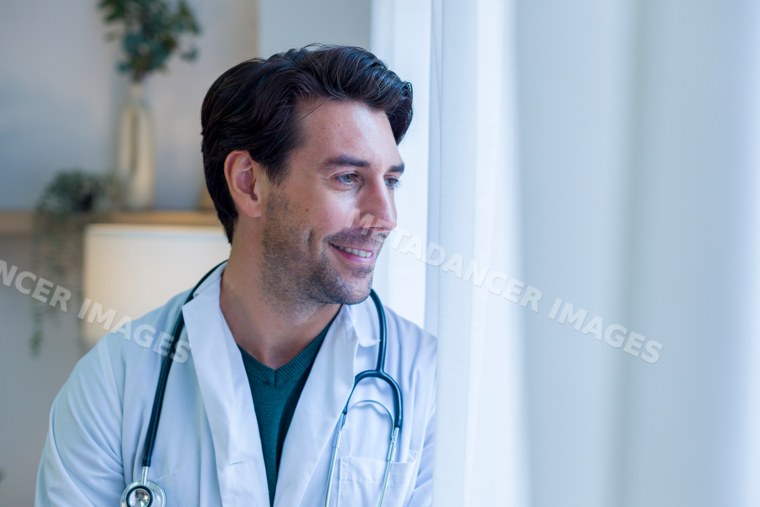 Doctor wearing white coat and stethoscope looking outside indoors