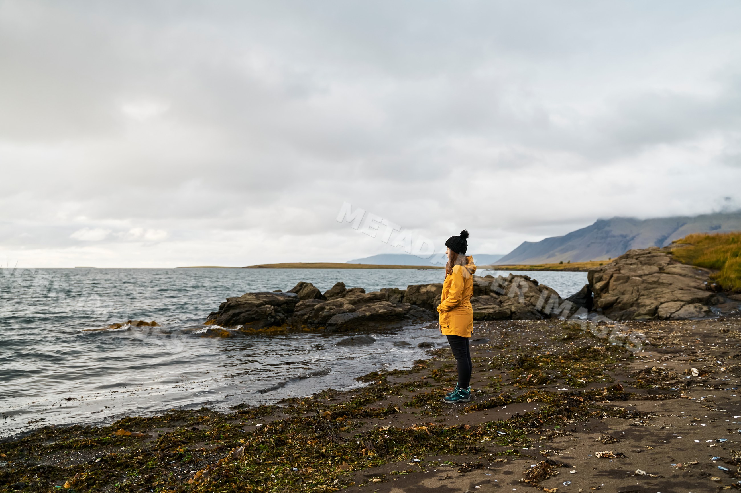A white woman in a yellow jacket stands on a rocky shoreline looking