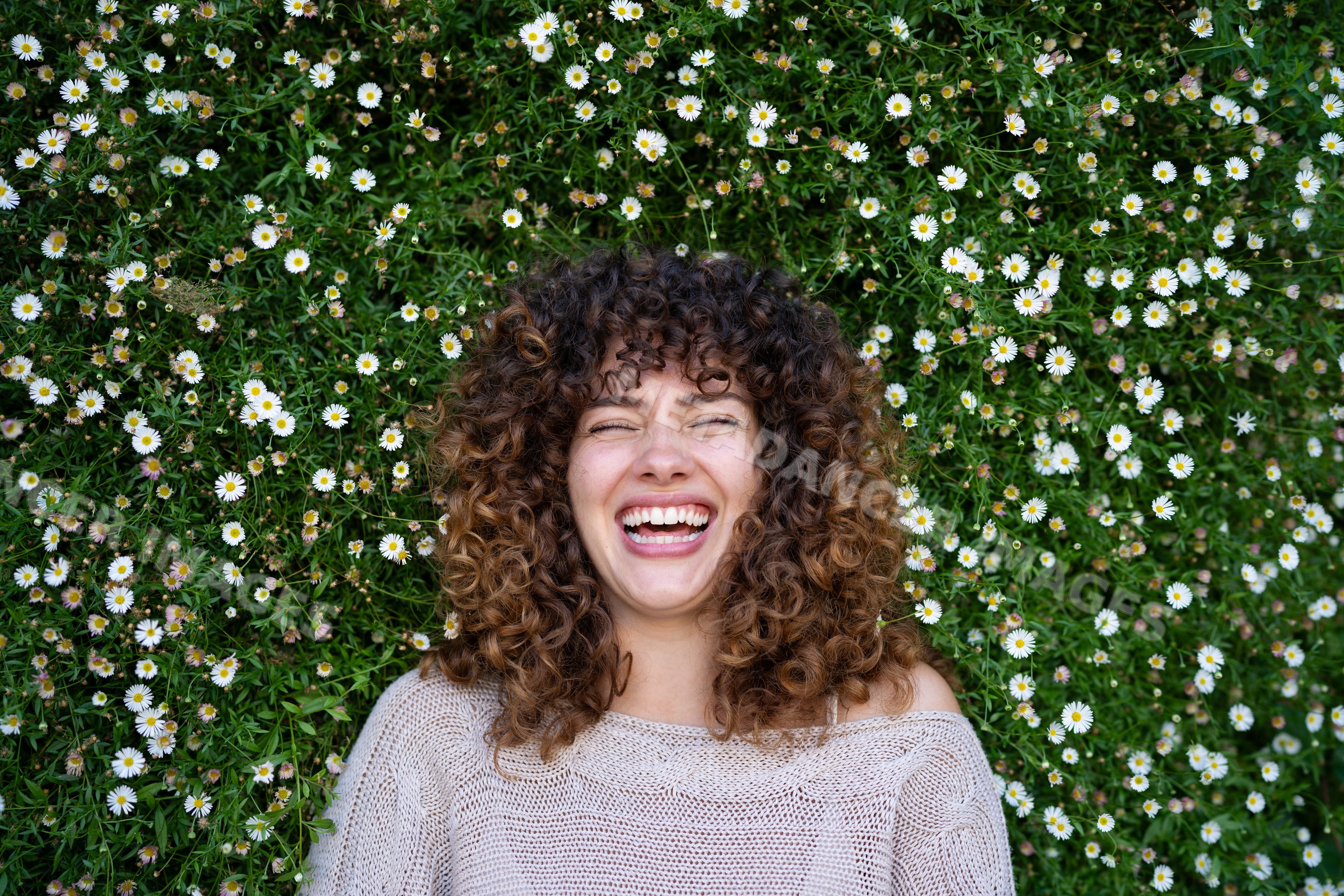 Woman with curly hair lies laughing in a field of white daisy flowers