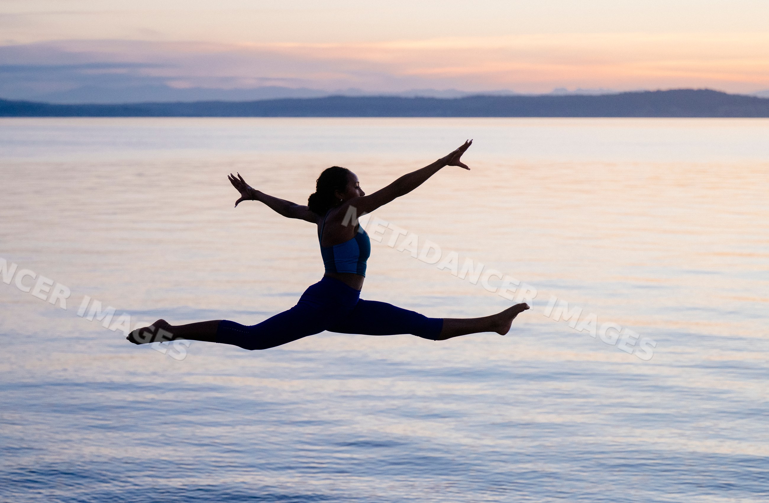 Hispanic woman performing jumping split yoga pose over lake at sunset