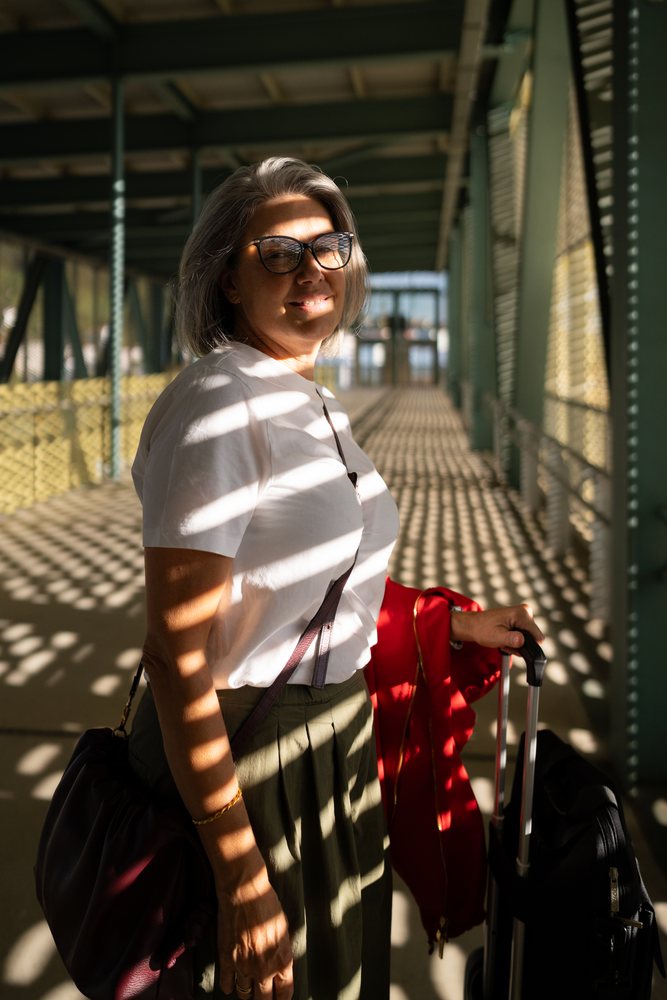 White woman standing in sunlit covered walkway with suitcase