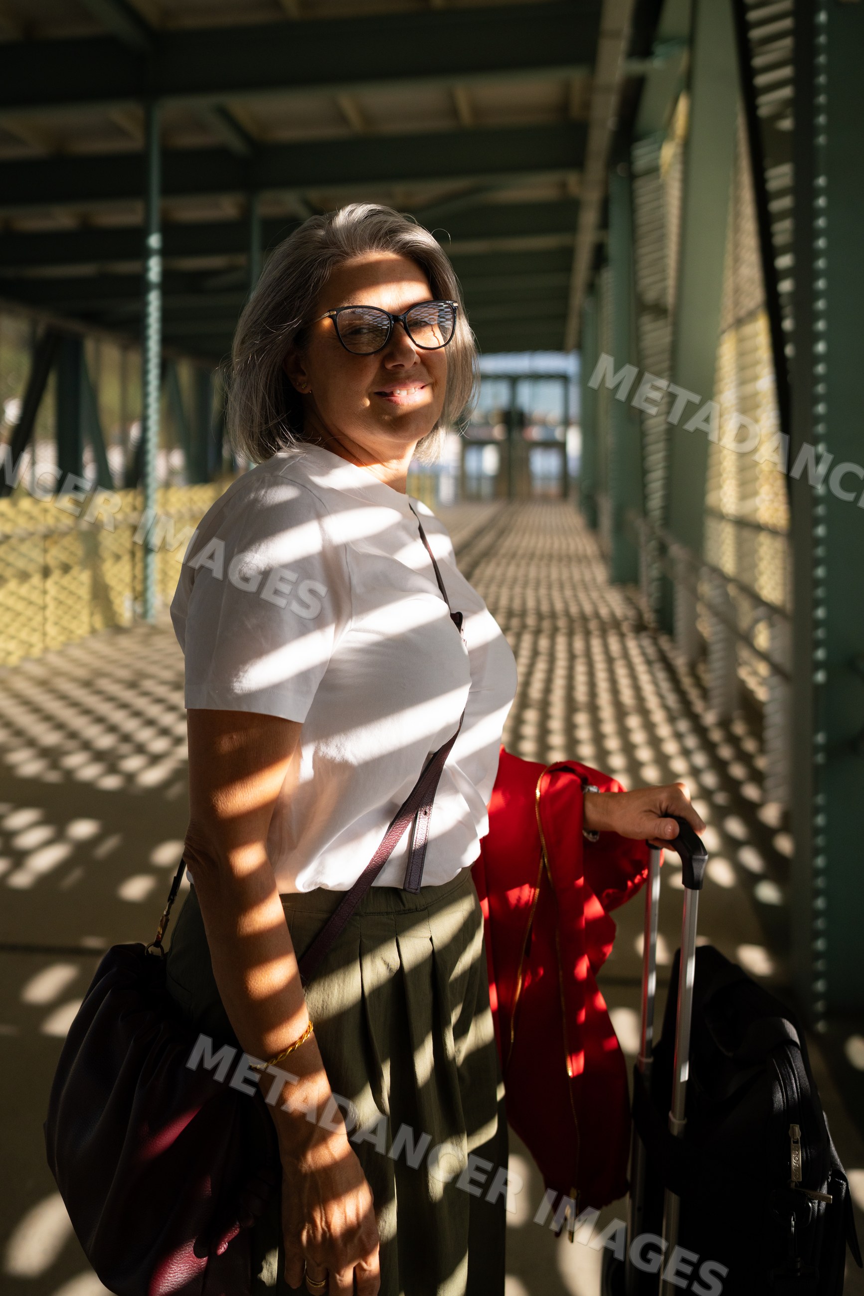 White woman standing in sunlit covered walkway with suitcase