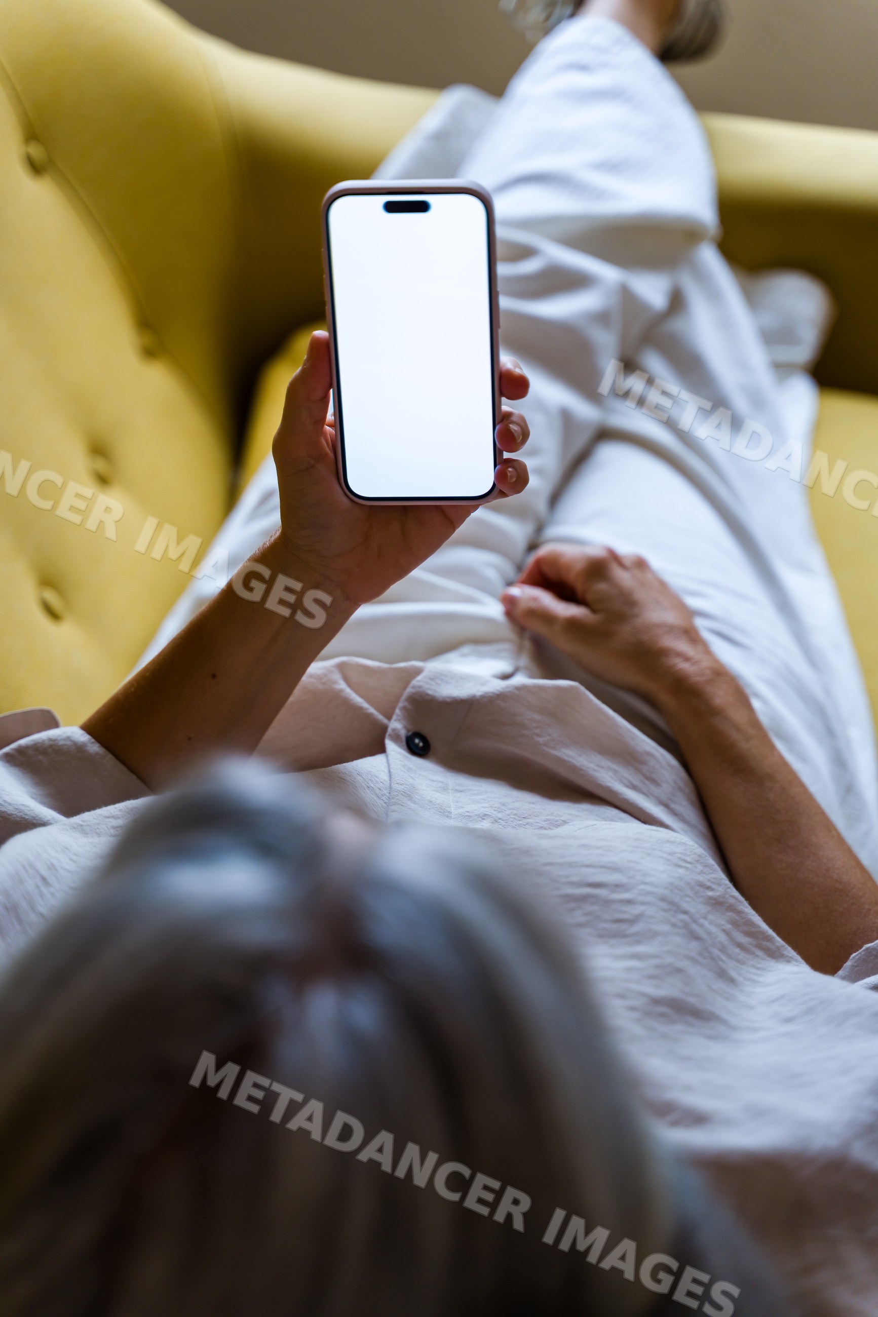 White person holding smartphone sitting on yellow sofa indoors