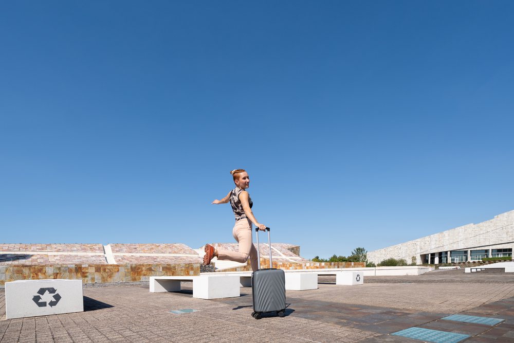 White woman jumping with suitcase in urban plaza with blue sky