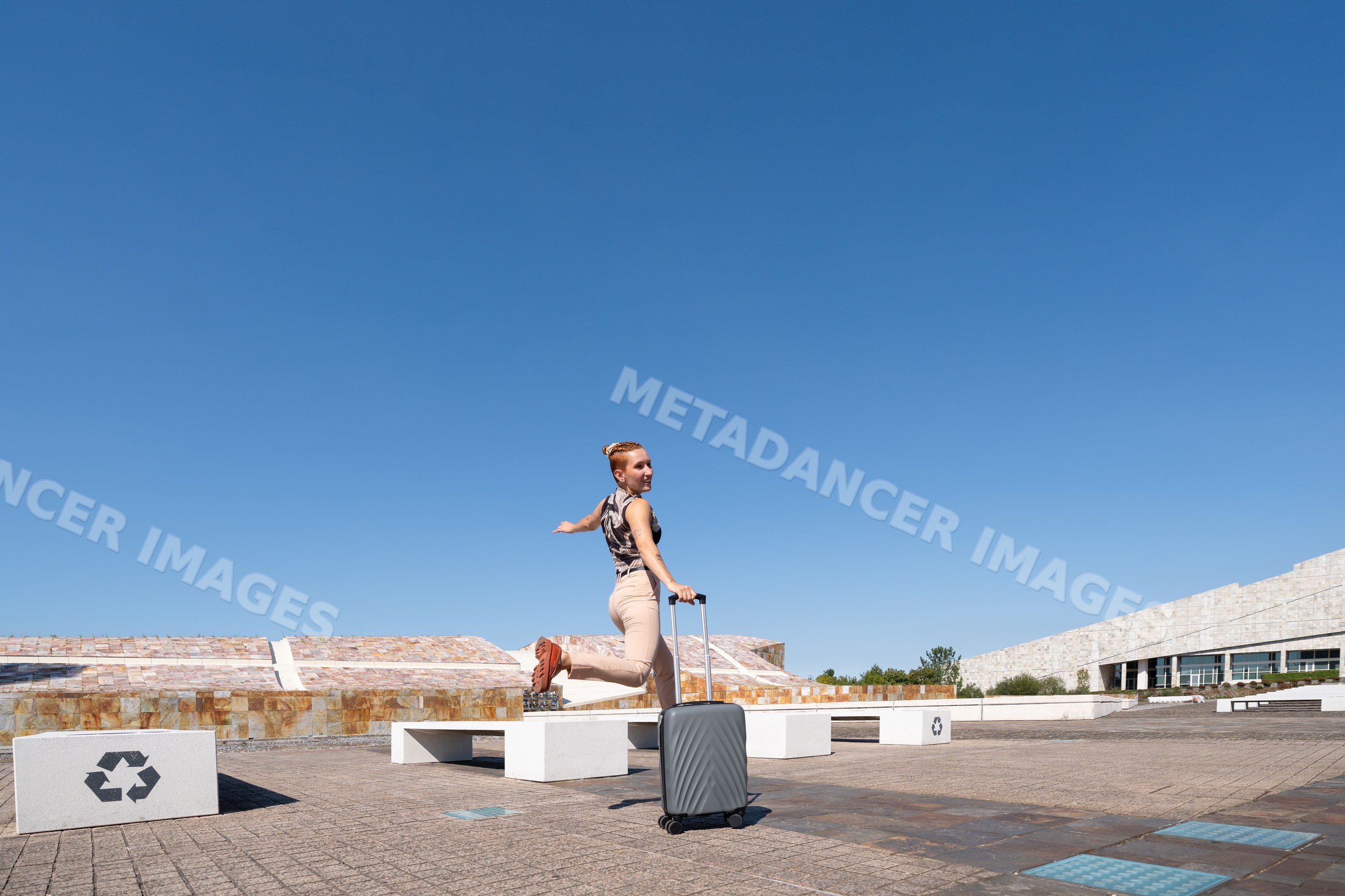 White woman jumping with suitcase in urban plaza with blue sky