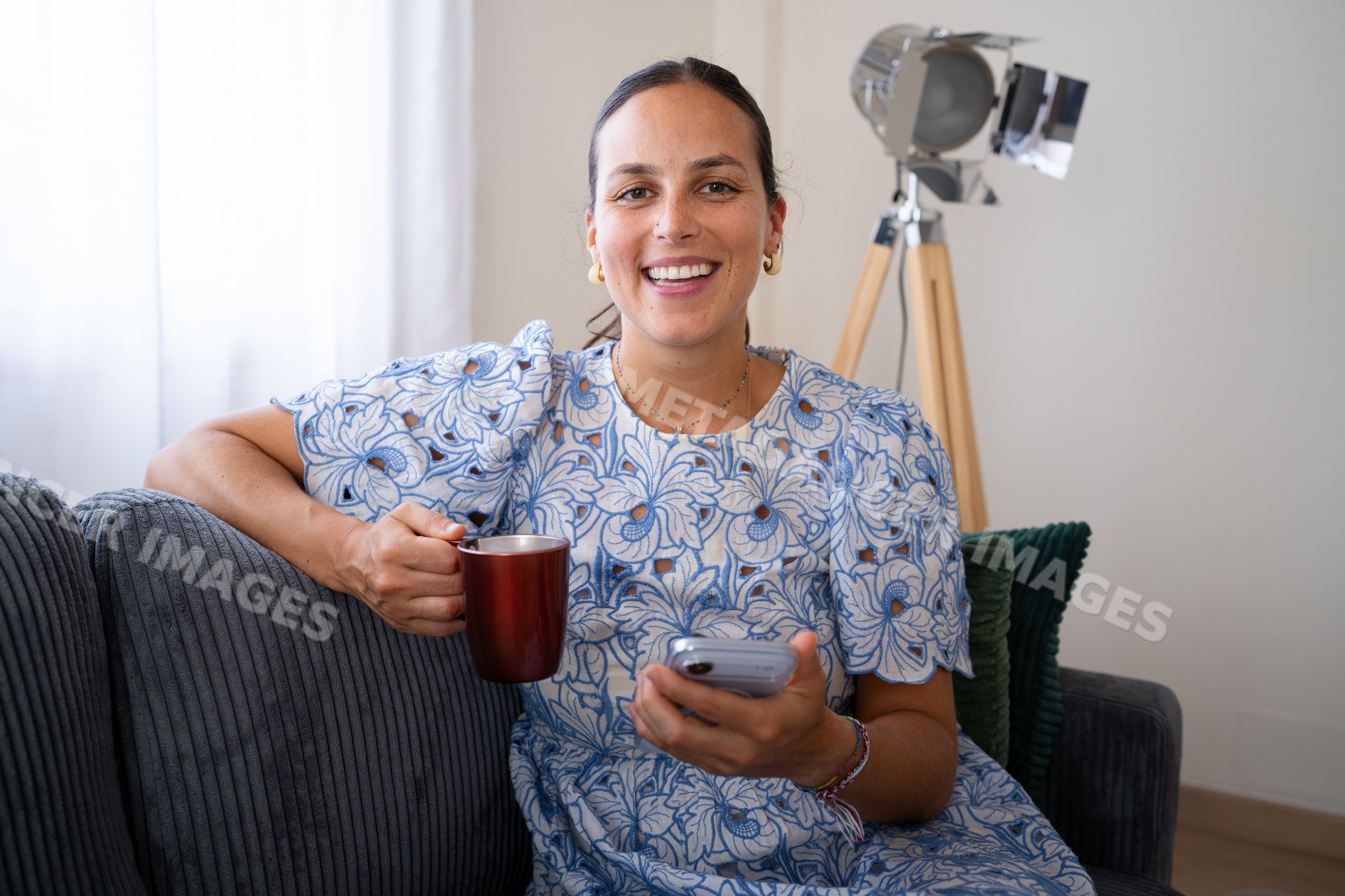 White woman smiling with mug and smartphone sitting on sofa indoors
