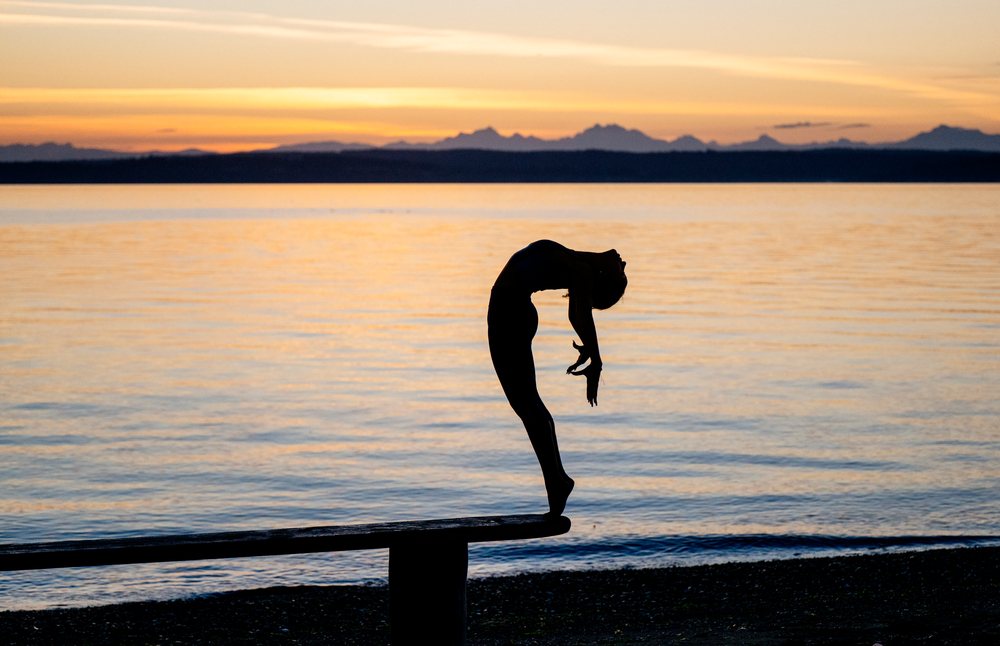 Hispanic woman performing backbend yoga pose on bench by water